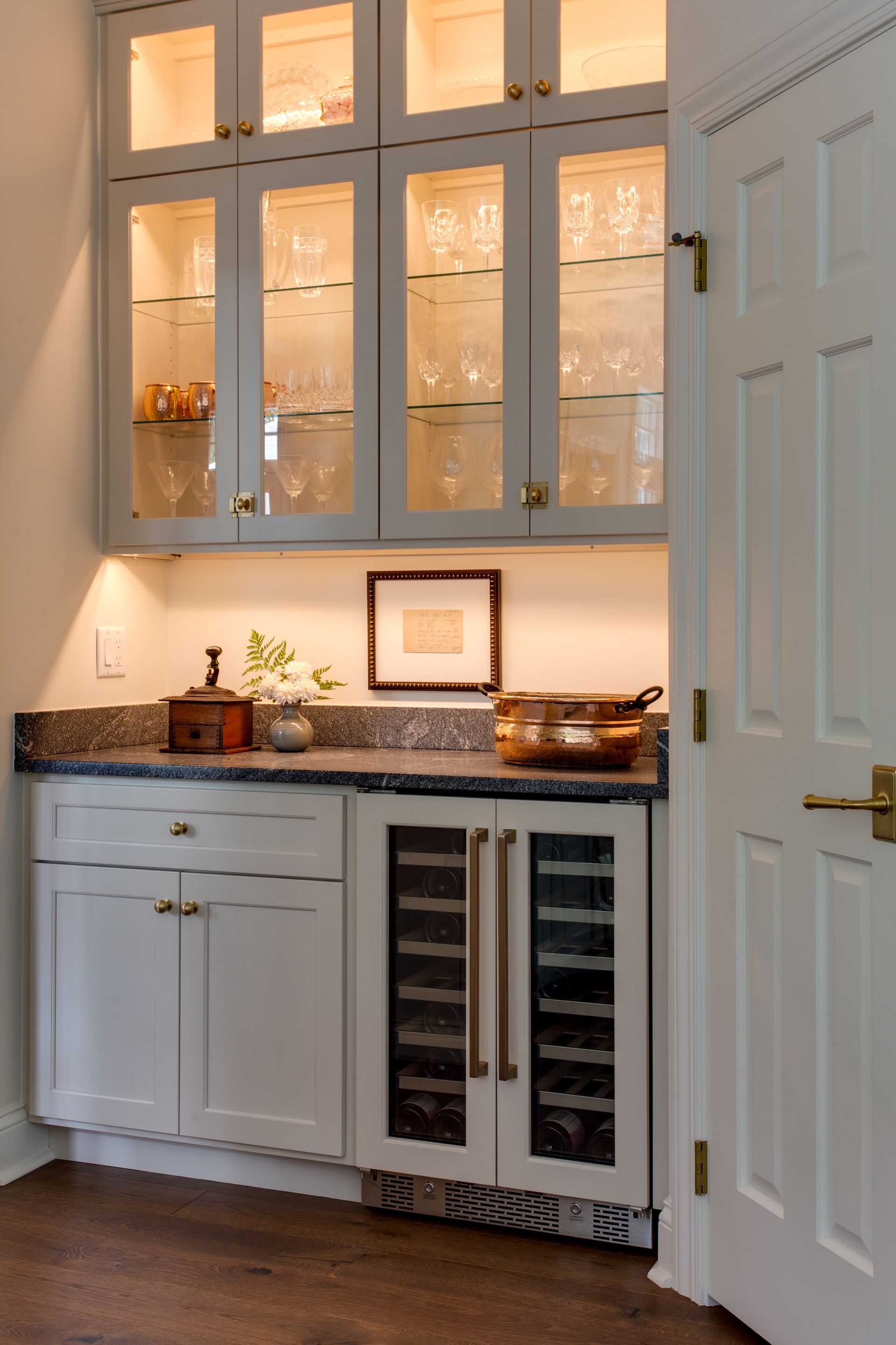 White built-in bar with glass-front cabinets, wine fridge, granite countertop, gold hardware, and a small framed artwork.