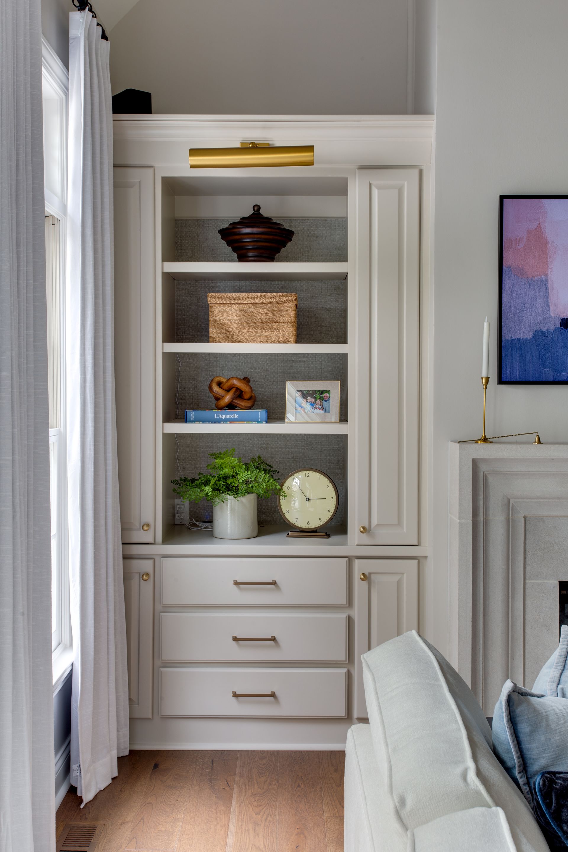 White built-in shelving unit with decor items. Includes drawers and a gold light fixture. Next to a window.