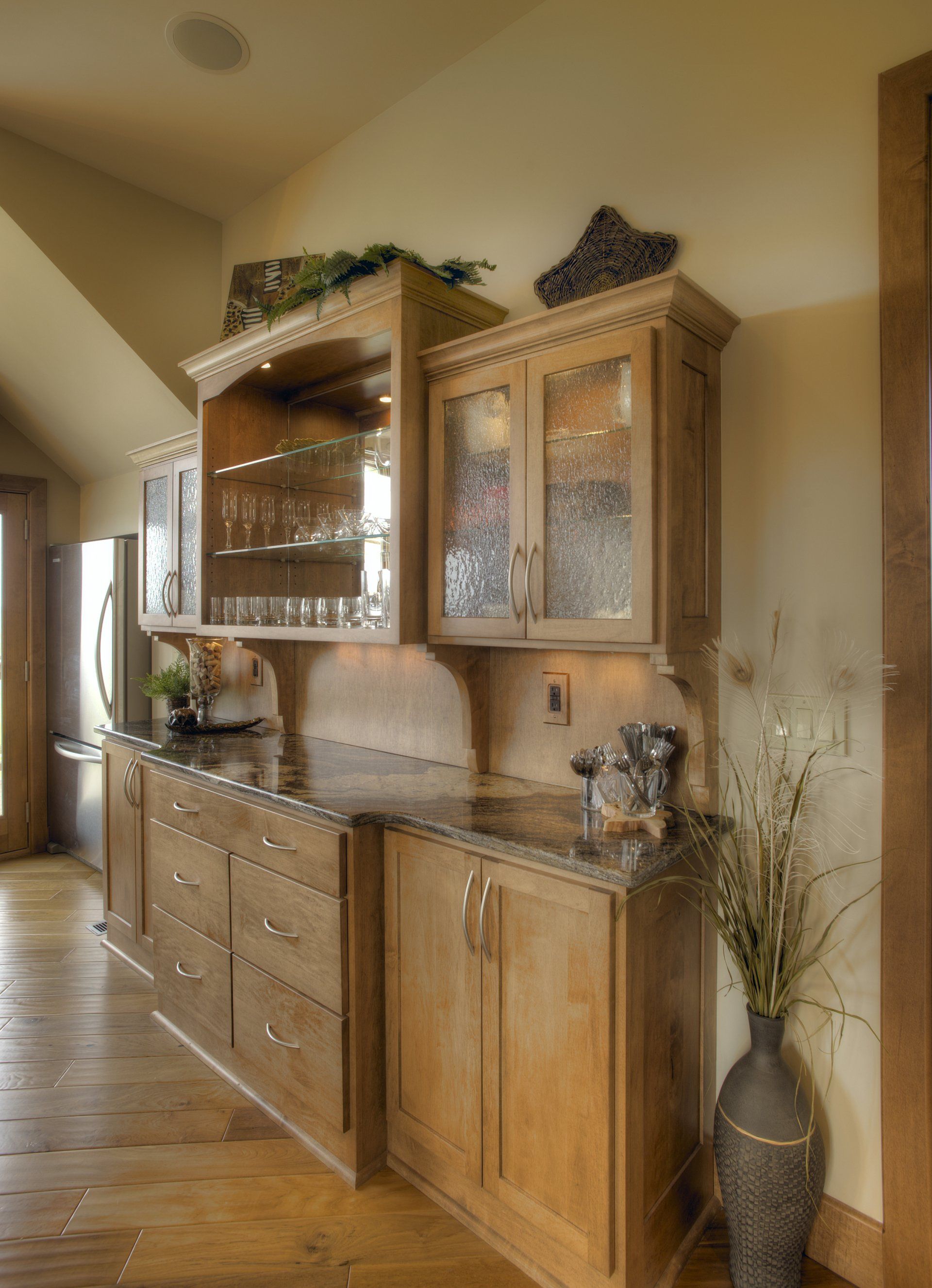 Wooden kitchen cabinets with granite countertops, glass-fronted upper cabinets, and a decorative vase.