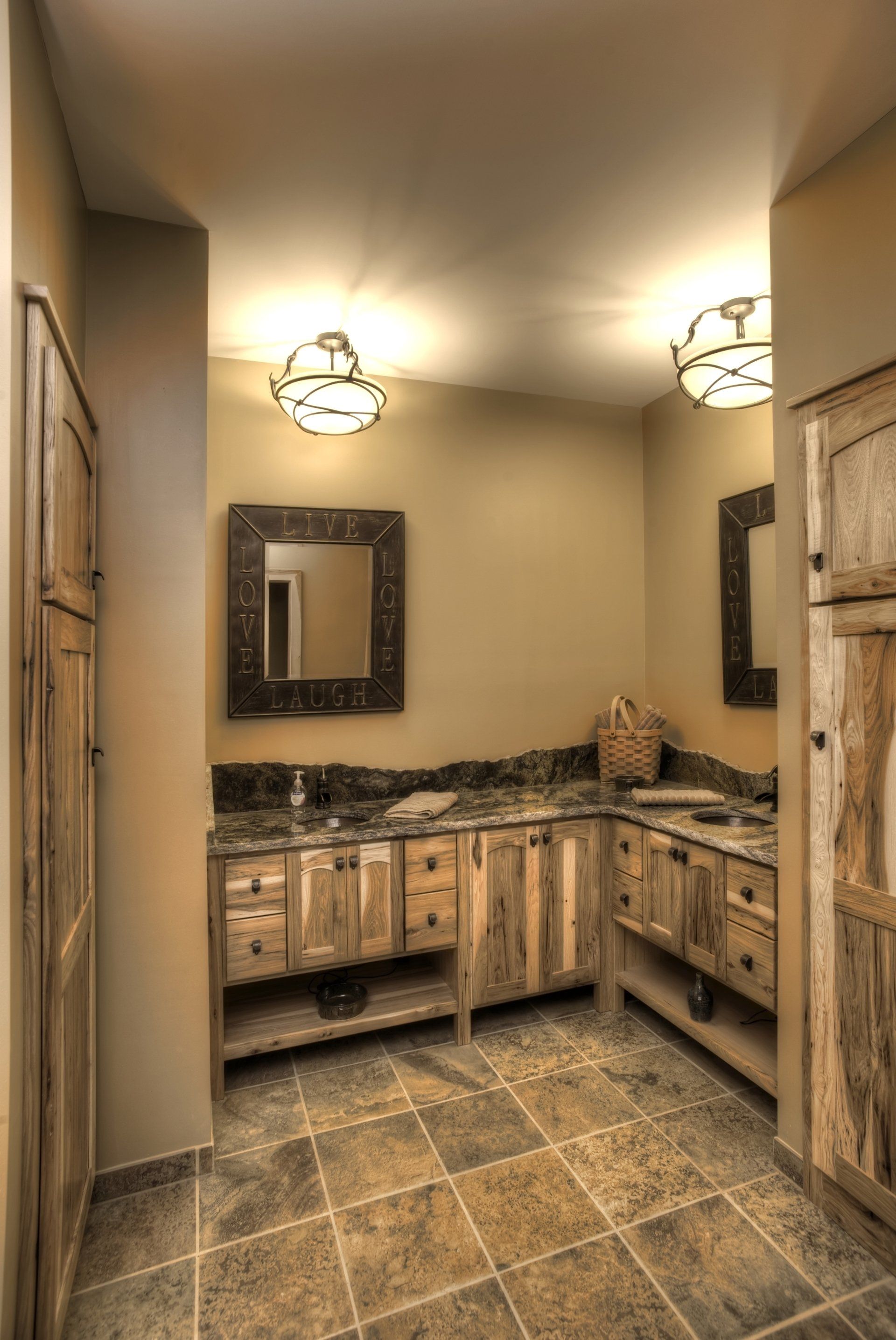 Bathroom with rustic wooden cabinetry, dark countertop, and tile floor, with two decorative light fixtures and mirrors.