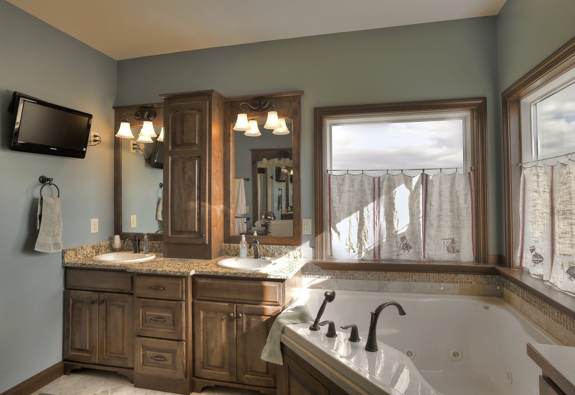 Bathroom with wood cabinets, dual sinks, soaking tub, and window with curtains.