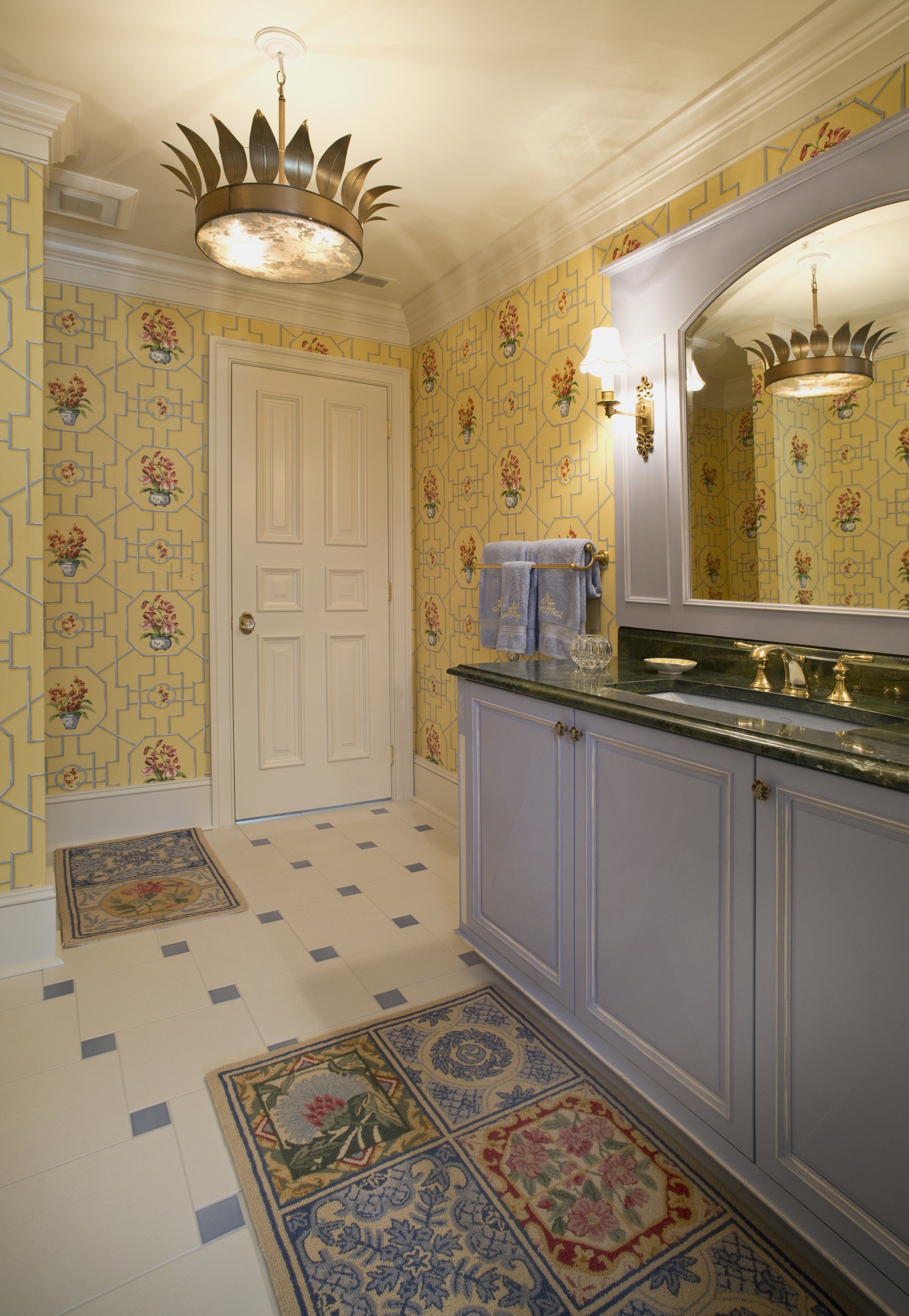 Bathroom with floral wallpaper, lavender vanity, crown-shaped light fixture, and patterned rugs on a tiled floor.