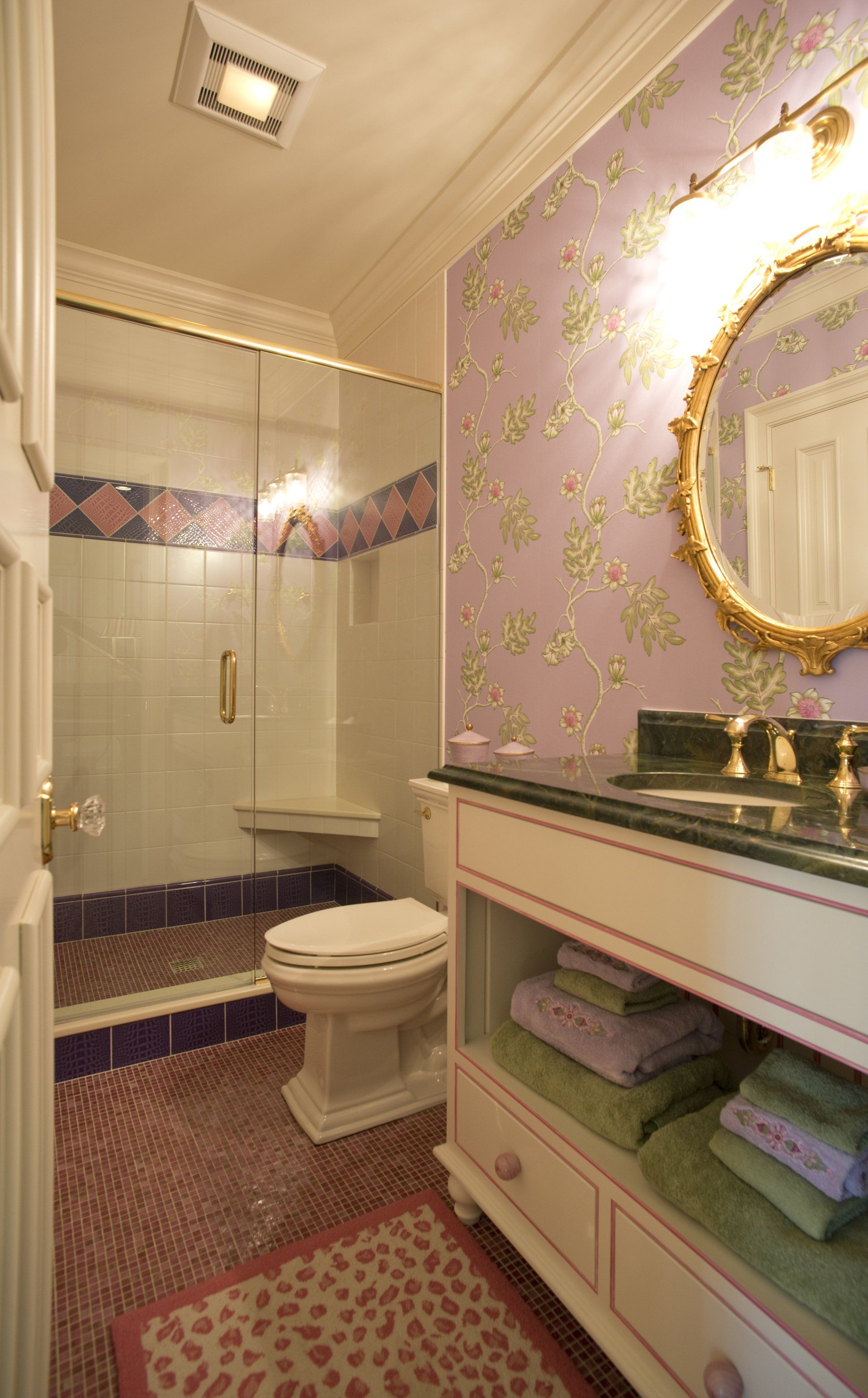 Bathroom with a patterned lavender and gold wallpaper, gold-framed mirror, and a white vanity with open storage.