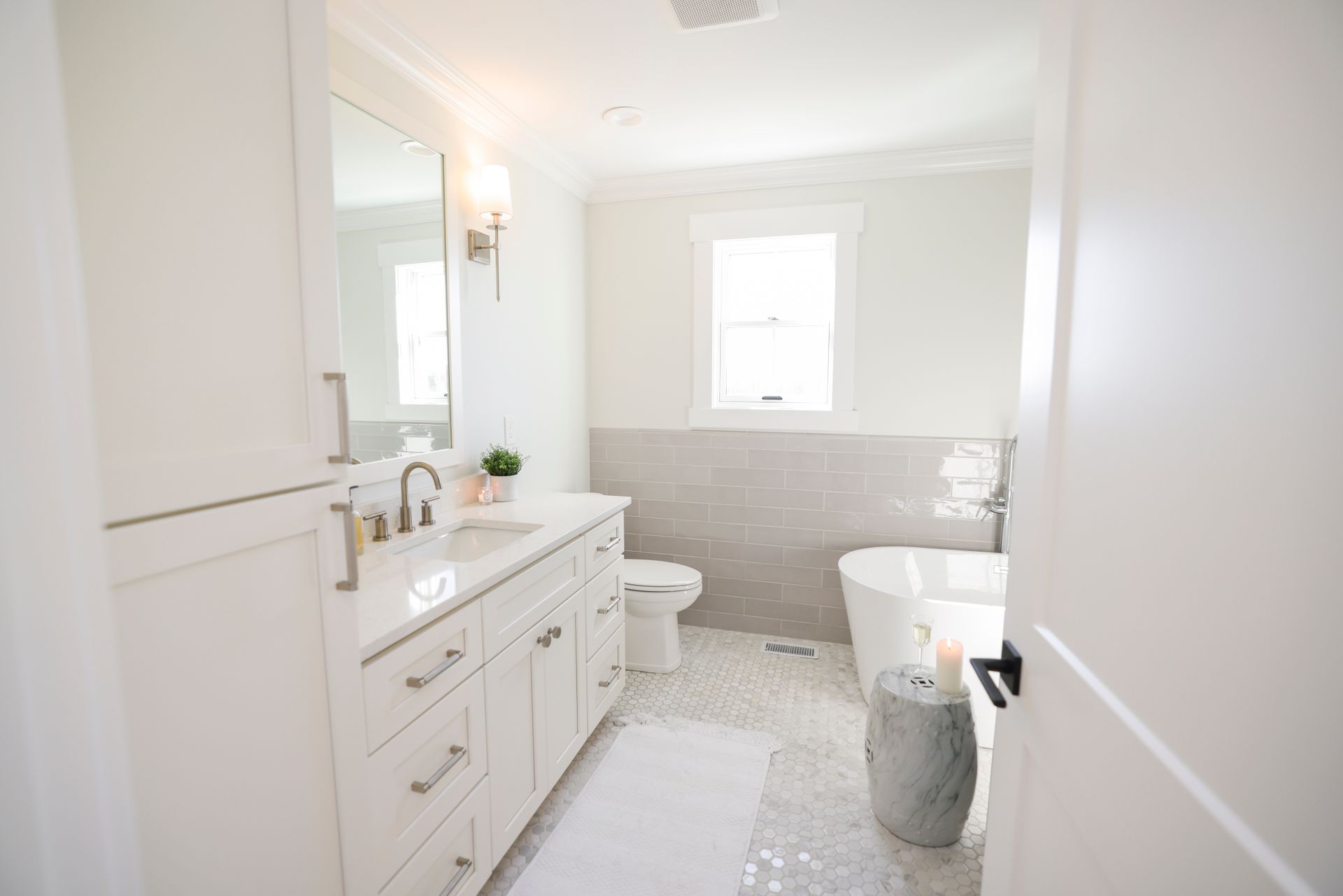 White bathroom with vanity, toilet, bathtub, and window. Pebble floor, light gray wall tile.