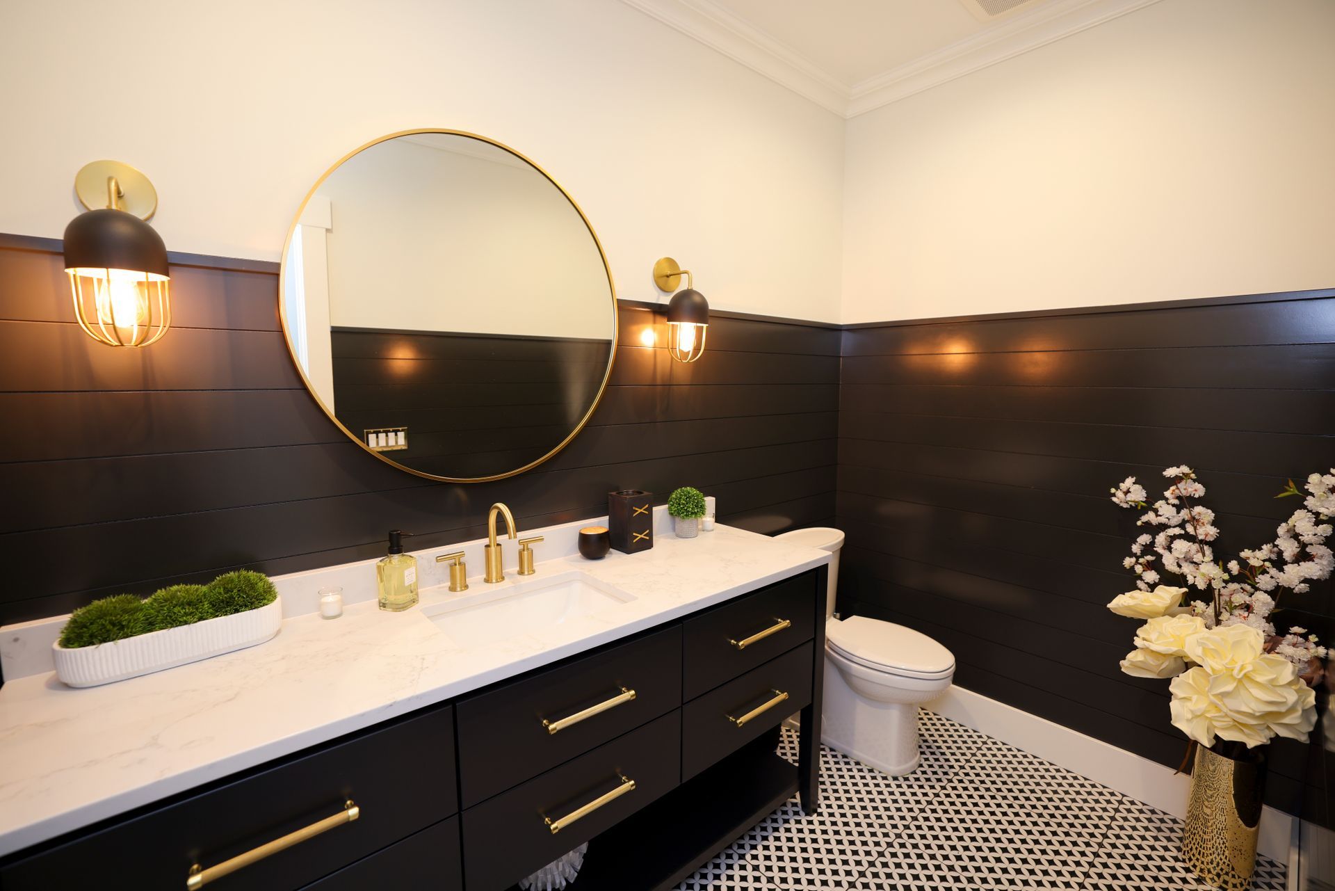 Modern black and white bathroom with gold accents: black vanity, round mirror, patterned floor, and sconces.