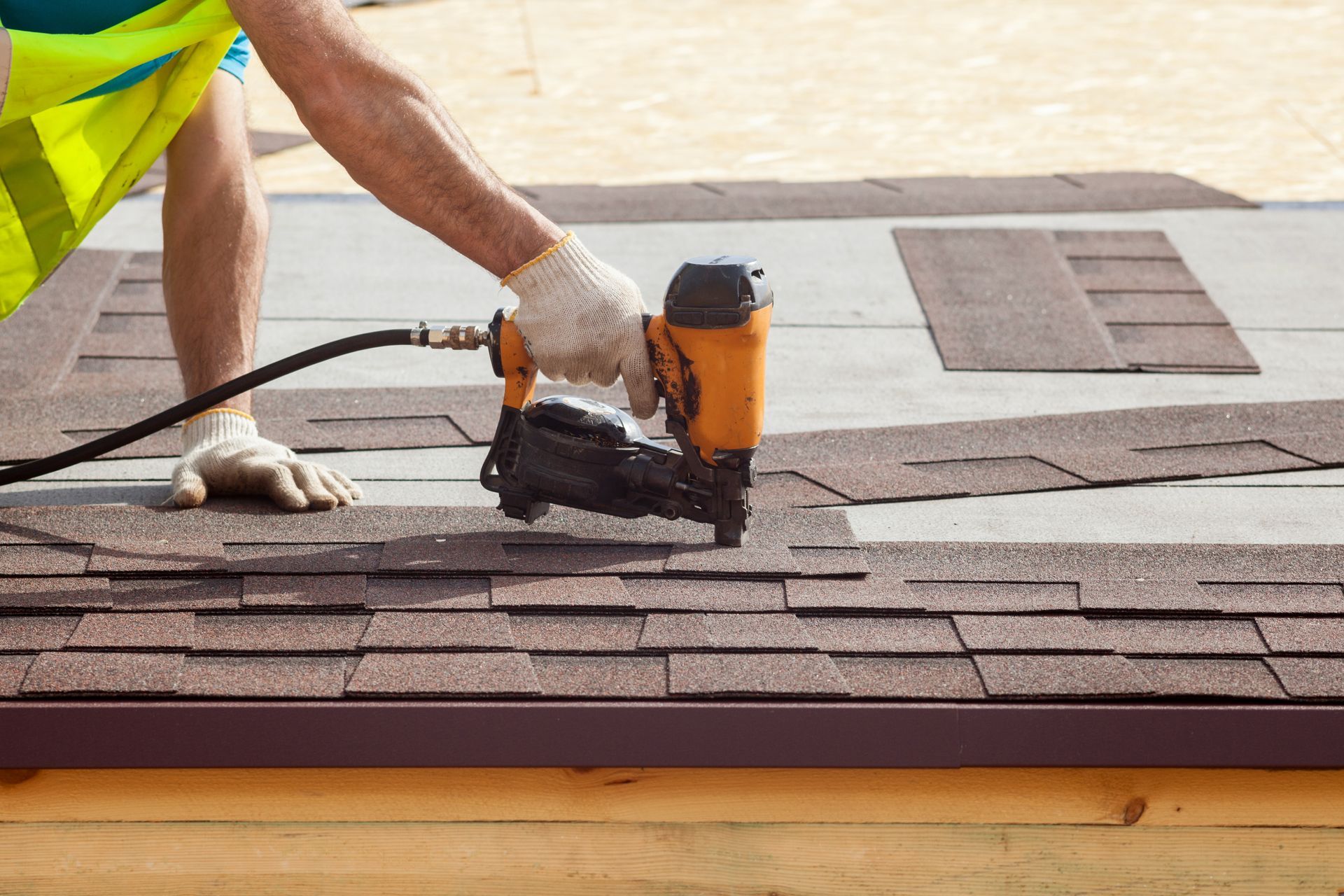 Worker installing asphalt shingles with a nail gun on a roof.