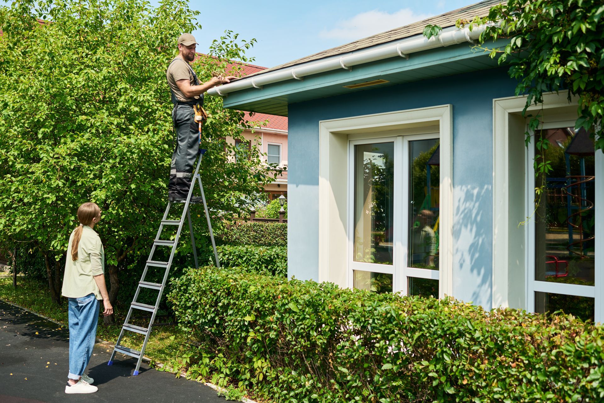 Contractor on a ladder repairing gutters while homeowner watches