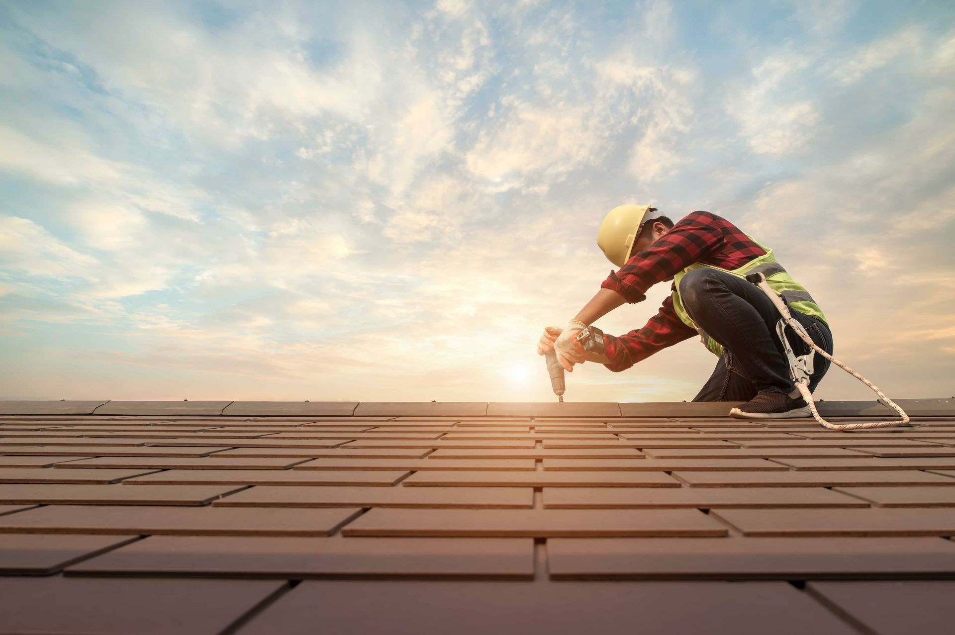 A roofing contractor installing shingles on a roof at sunset using power tools and safety gear.