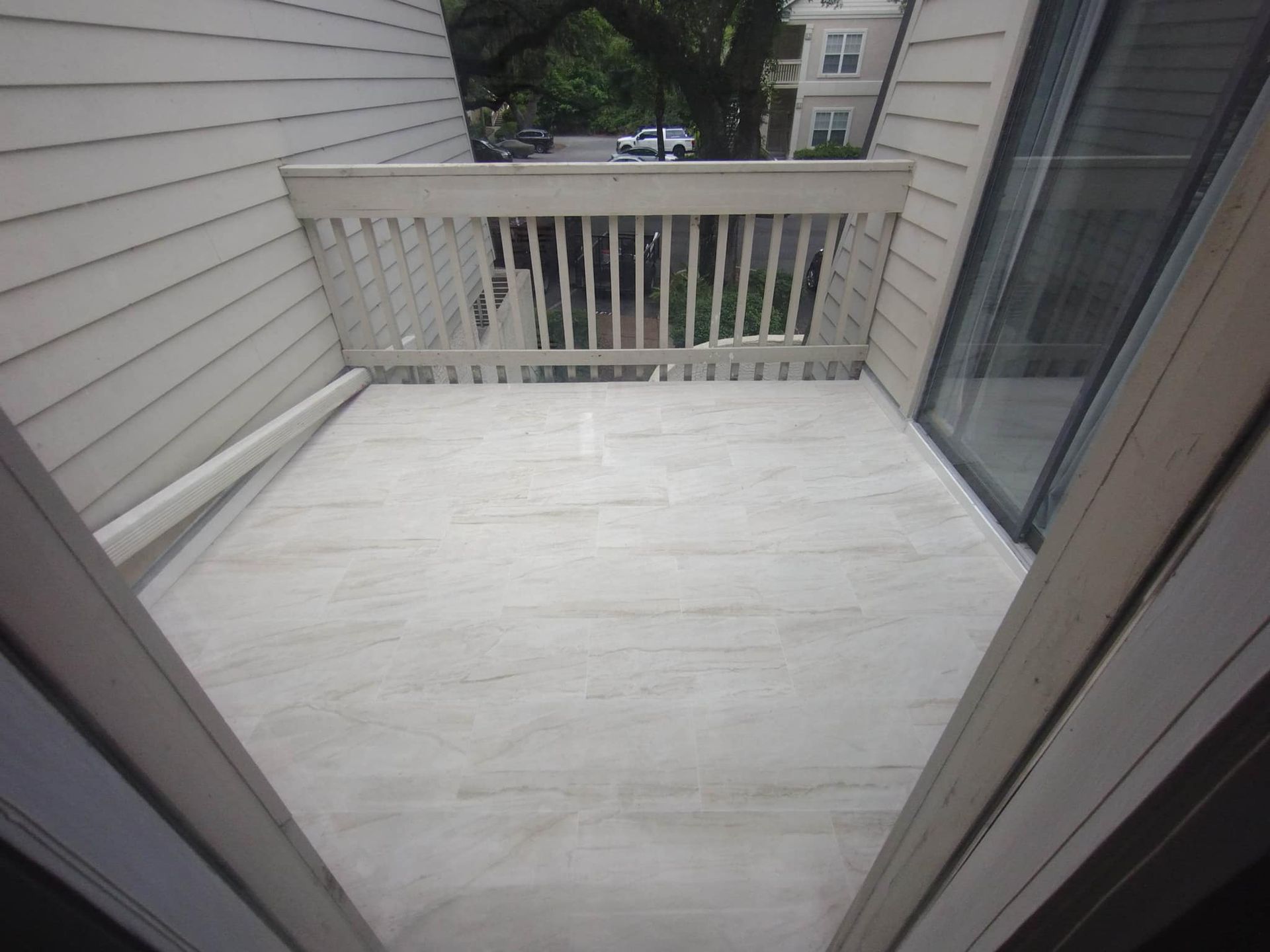 A balcony with light-colored flooring, beige siding, and a wooden railing overlooking trees and a distant building.