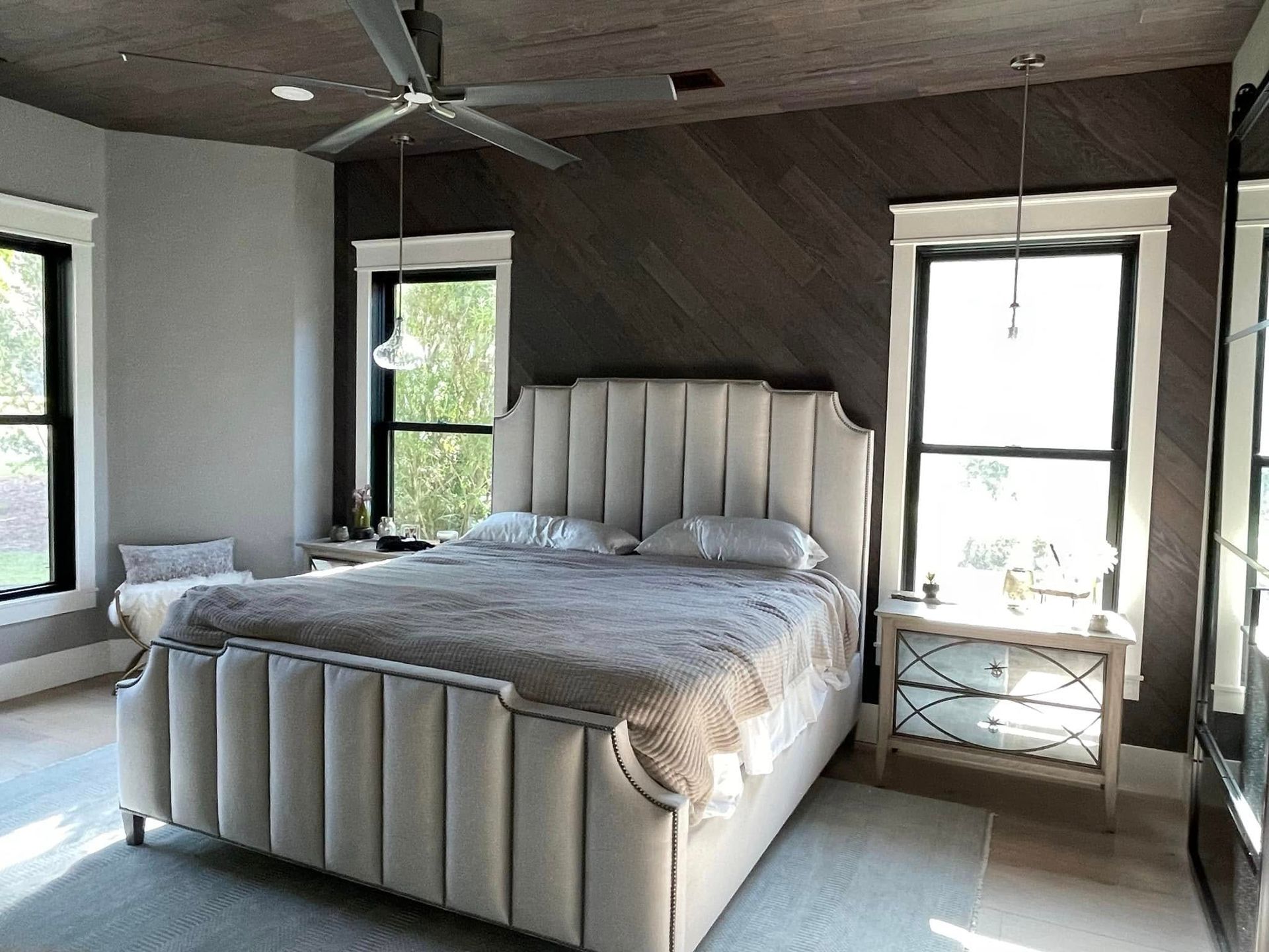 A bright bedroom featuring a tall, cream-colored, tufted headboard, dark wood paneled walls, and two bedside tables.