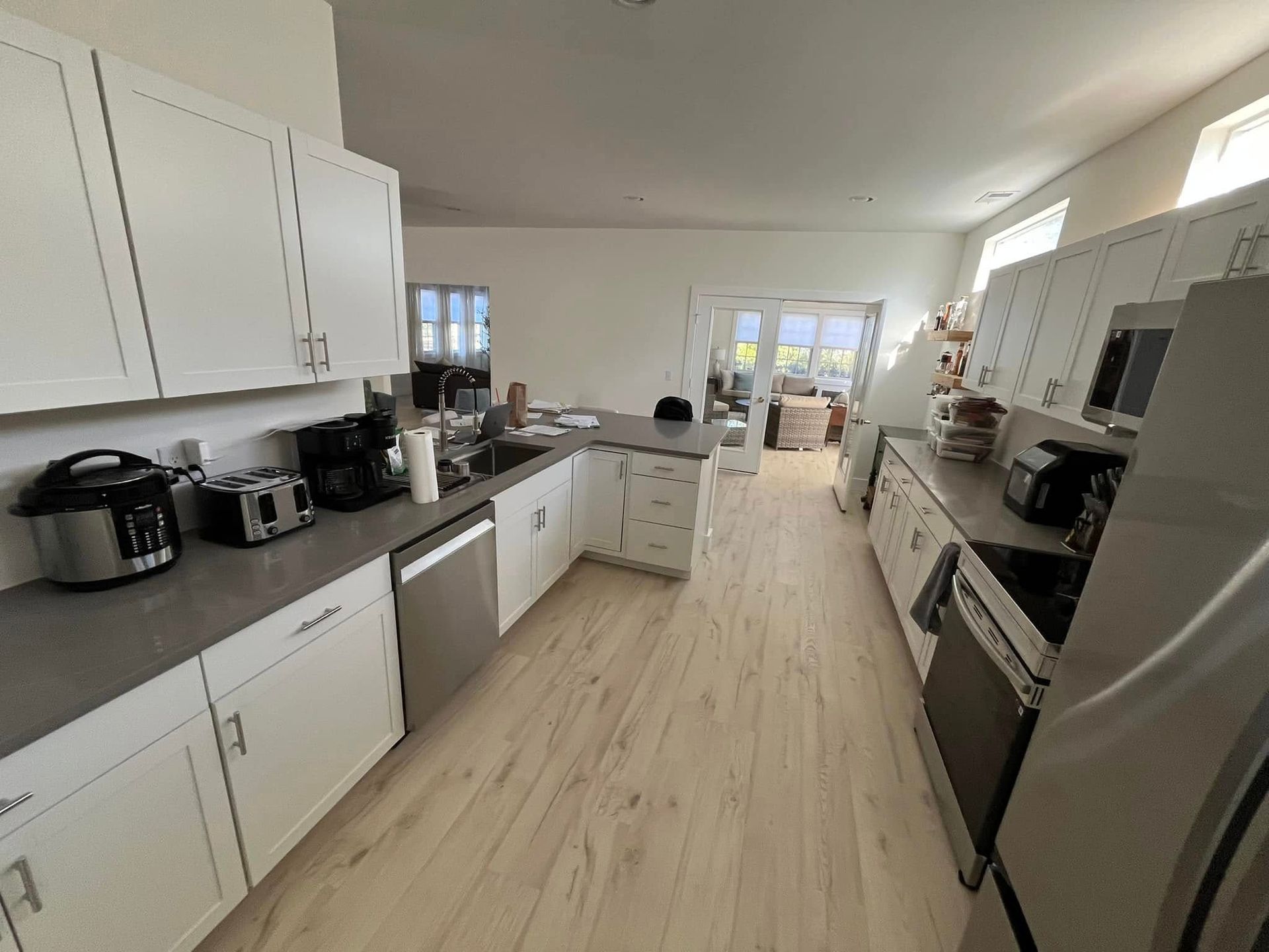 A bright, modern kitchen features white cabinets, grey countertops, stainless steel appliances, and light wood flooring.
