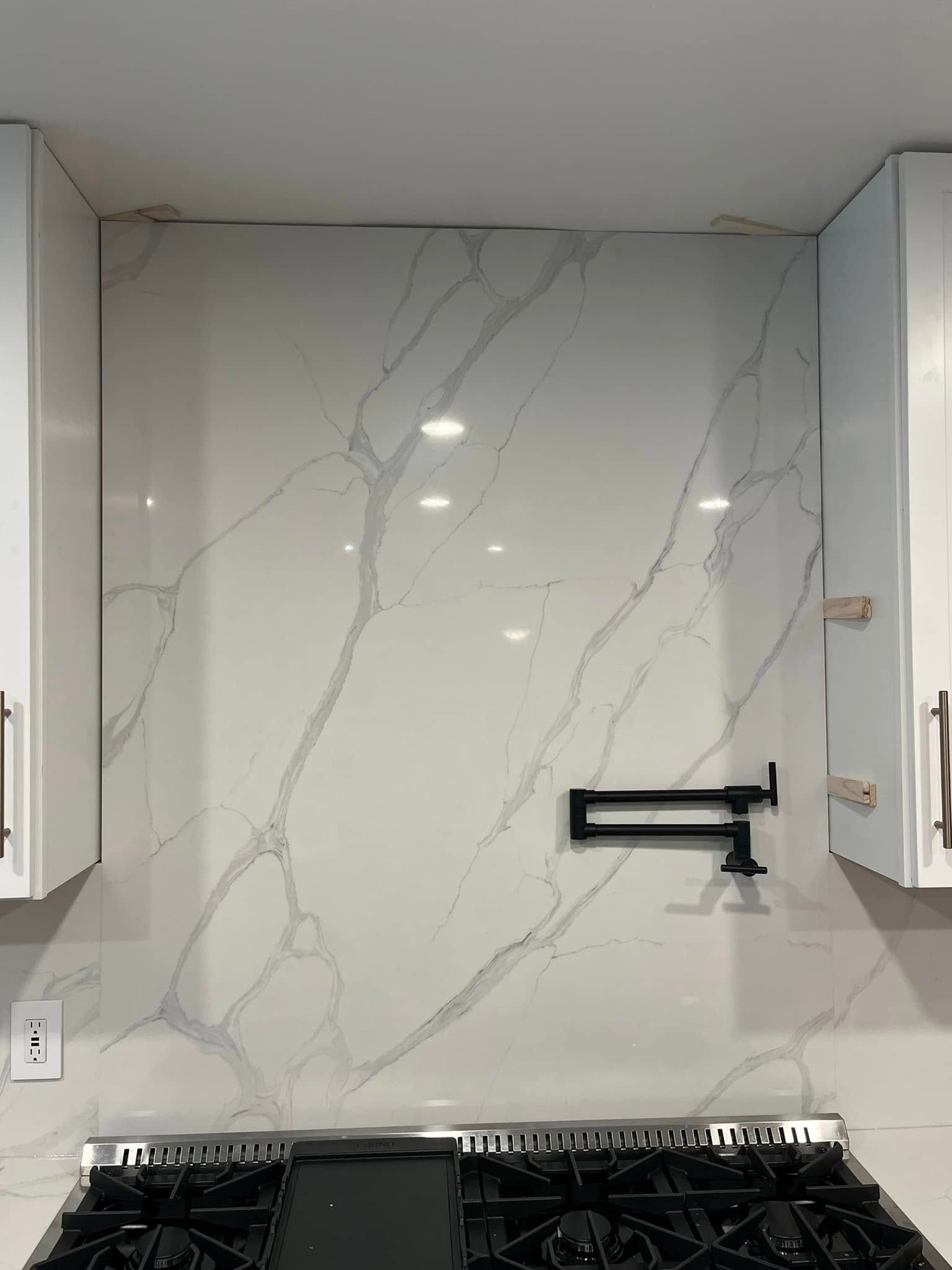 A marble-look kitchen backsplash with a mounted black pot filler faucet above a gas stove, flanked by white cabinets.
