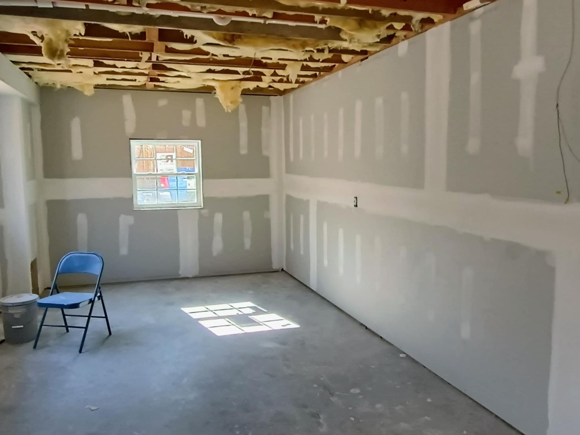 An unfinished room with gray drywall, exposed ceiling insulation, a small window, and a lone blue folding chair.
