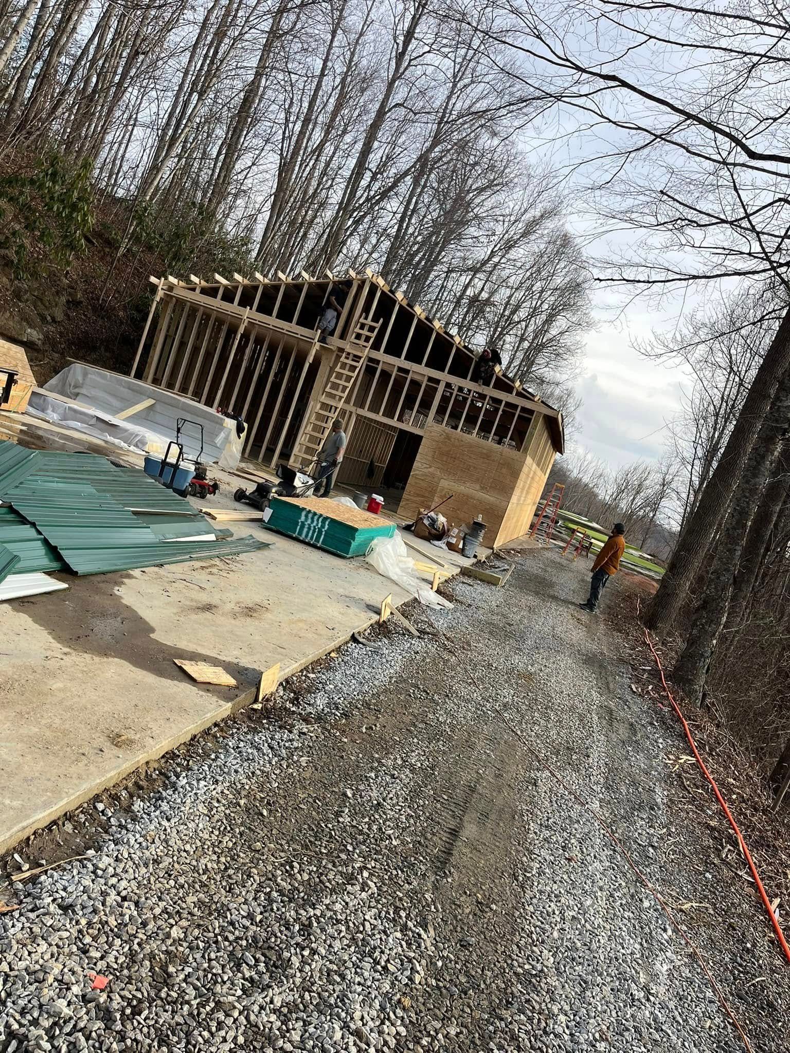 A wooden building frame under construction near a gravel path and trees, with a person standing to the right.