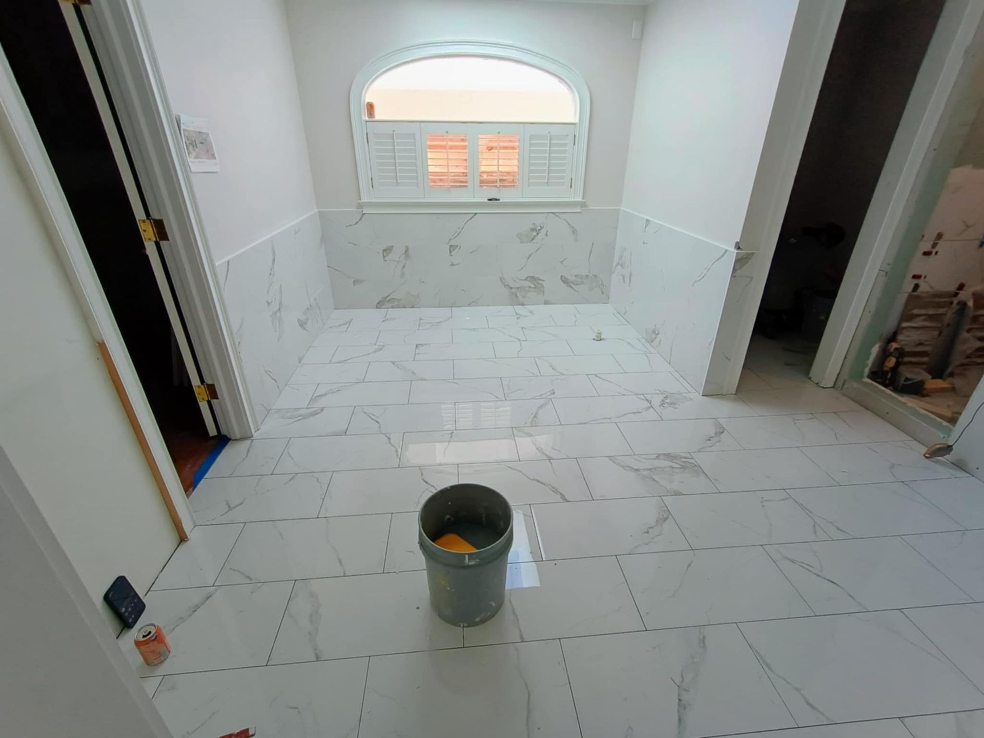 A renovated room with white marble-patterned tiles on the floor and lower walls, featuring a large arched window.