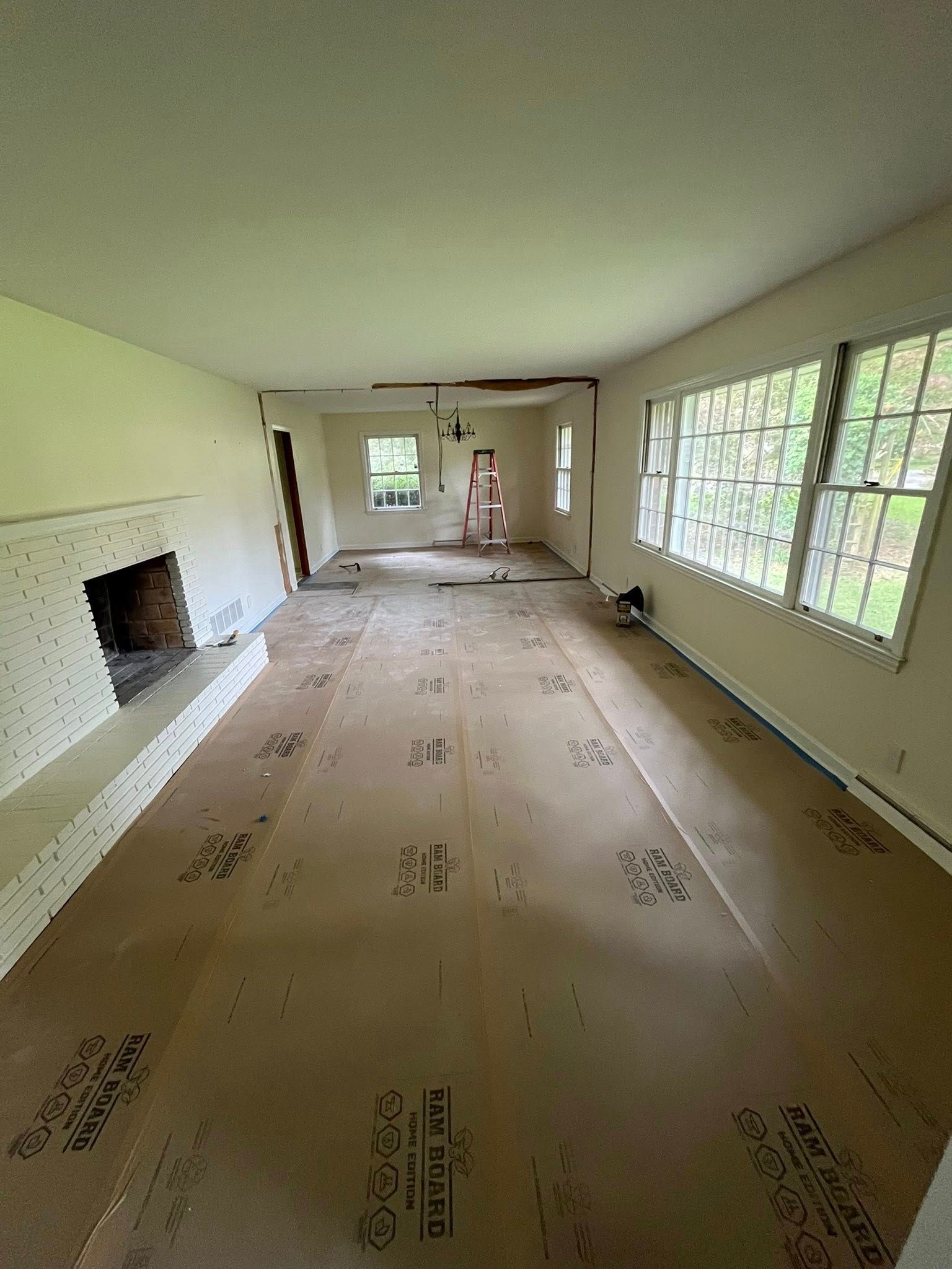 A long, empty room undergoing renovation, featuring paper-covered floors, a fireplace, and large windows.