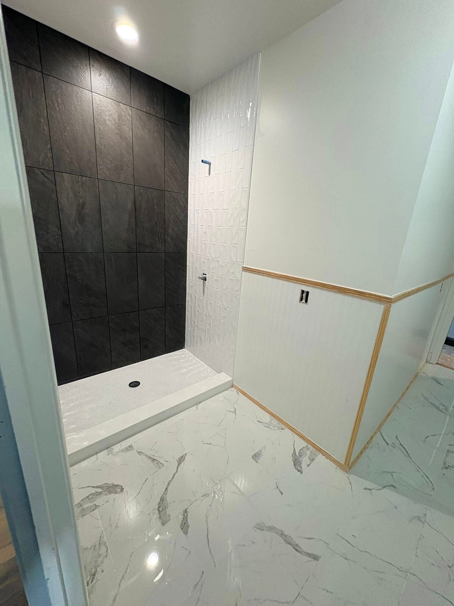 A modern bathroom featuring a shower with black tile, white marble floors, and off-white walls with a decorative trim.