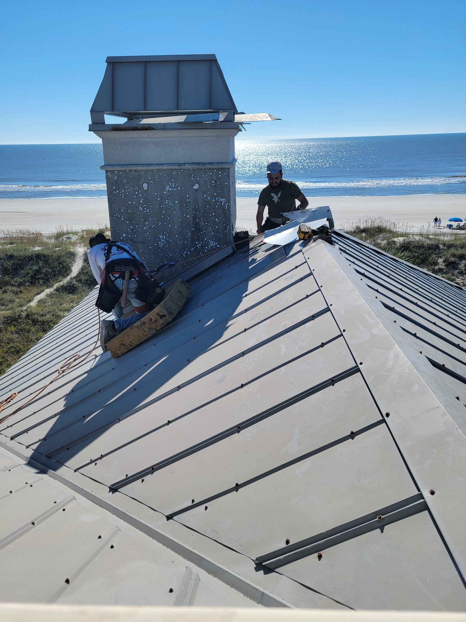 Two workers perform repairs on a metal roof near a chimney, with the ocean and beach visible in the background.