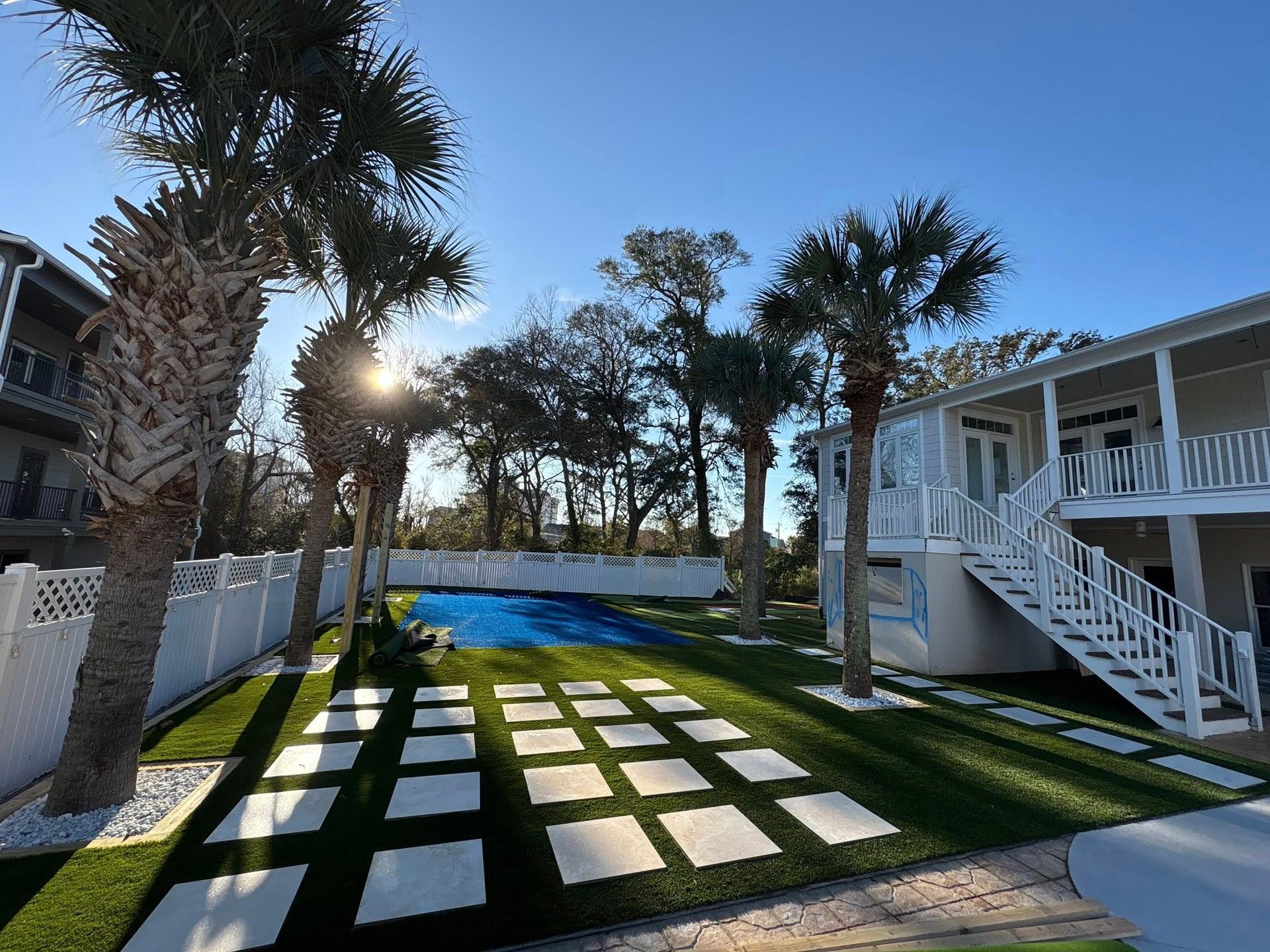 A yard with square stone pavers set in artificial grass leading to a bright blue pool beside a two-story house.