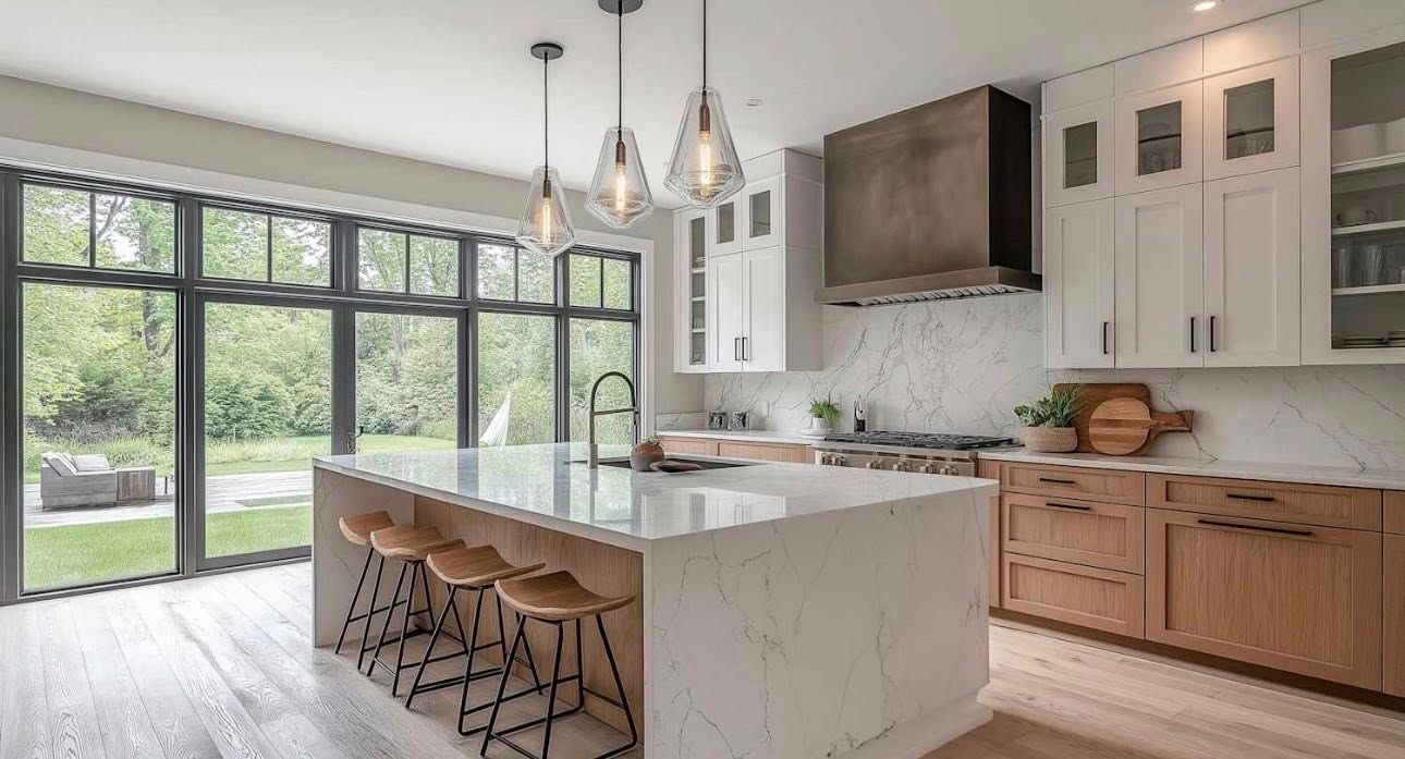 A modern kitchen with a large marble island, wood and white cabinets, a bronze range hood, and large windows to a yard.