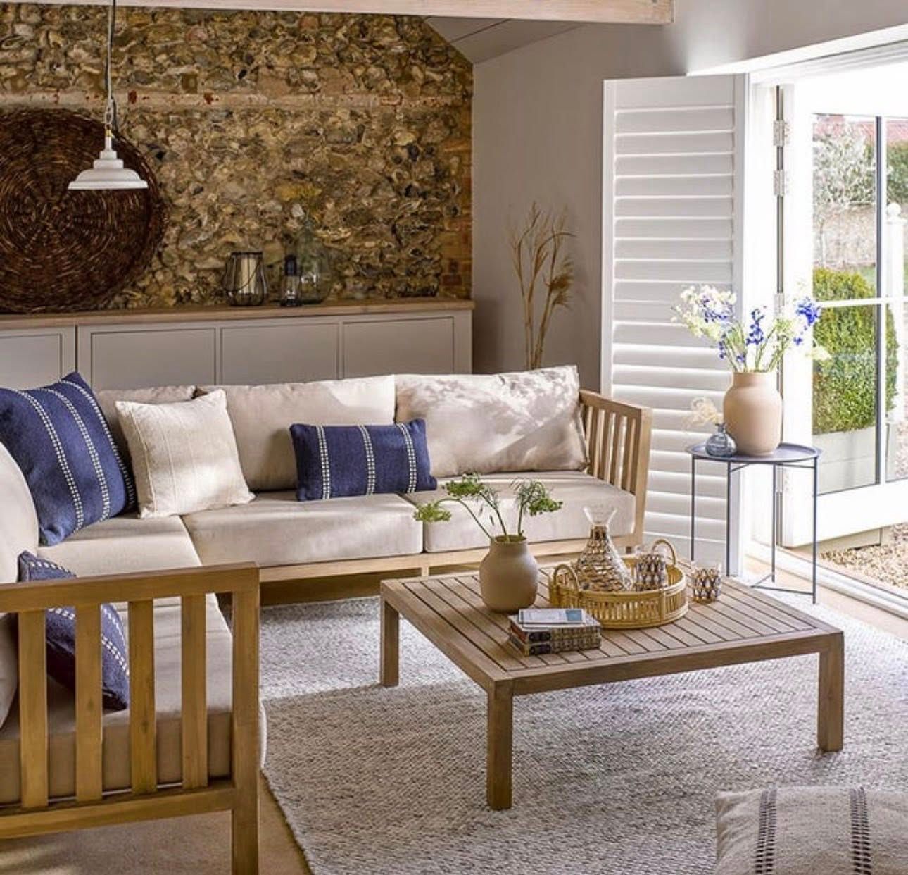 A light wood sofa with beige cushions and blue accent pillows sits on a white rug in a room with a textured stone wall.