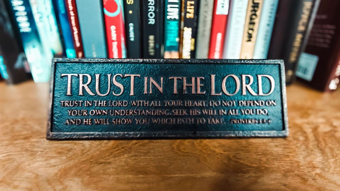 A sign that says `` trust in the lord '' is sitting on a wooden table in front of a bookshelf.