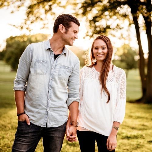 Couple holding hands smiling outdoors; man in denim shirt, woman in white top.