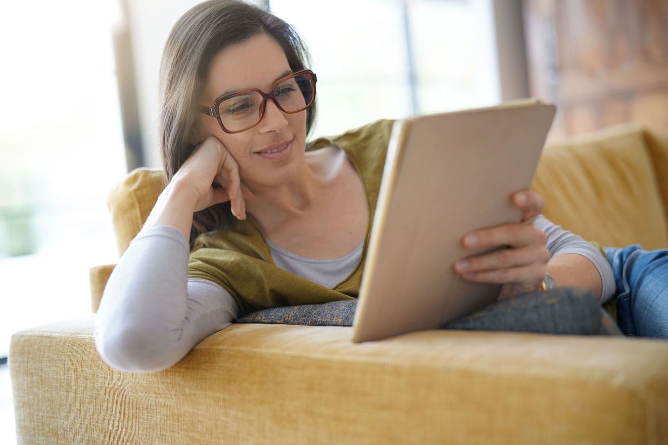 Woman reclining on yellow couch, wearing glasses, reading tablet.