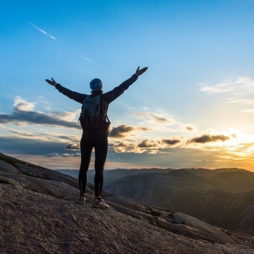 Person with arms raised on a mountaintop, overlooking a valley at sunset.