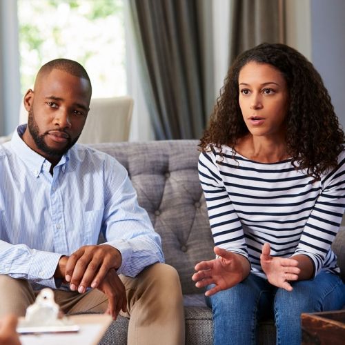 Couple in therapy session, woman gesturing, man looking on, seated on couch.