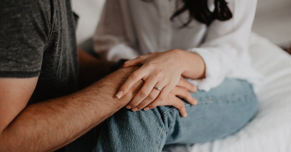 Couple holding hands, one wearing engagement ring. Man in gray shirt, woman in white shirt and jeans.