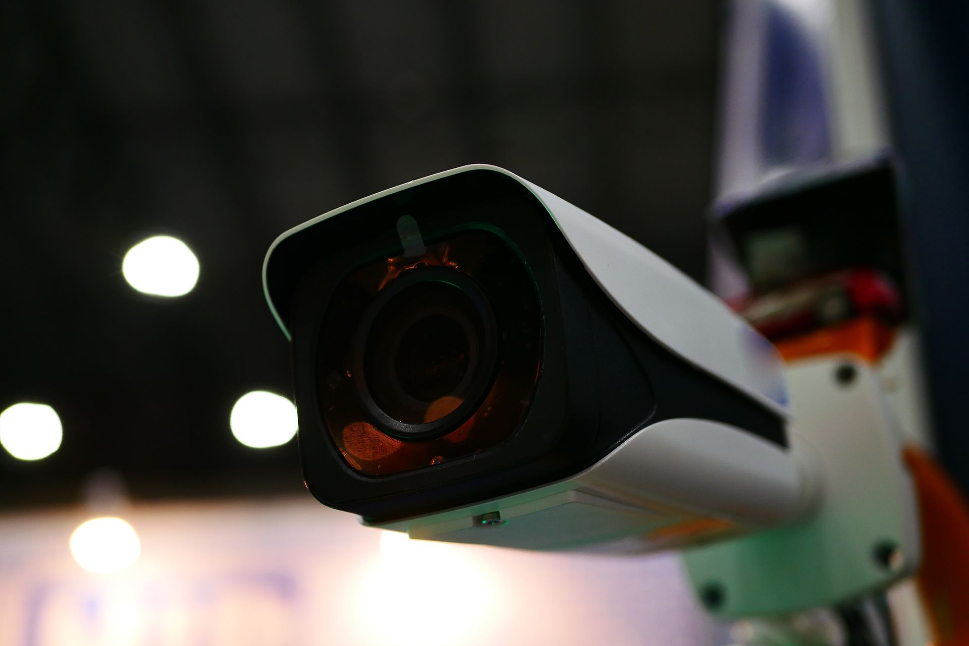 Close-up of a security camera with a black lens and white body, mounted on an orange and blue pole indoors. Close-up of a security camera with a black lens and white body, mounted on an orange and blue pole indoors.