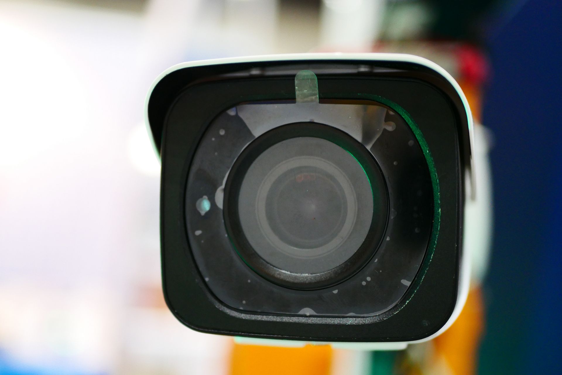 Close-up of a security camera with a black lens and white body, mounted on an orange and blue pole indoors. Close-up of a security camera with a black lens and white body, mounted on an orange and blue pole indoors.