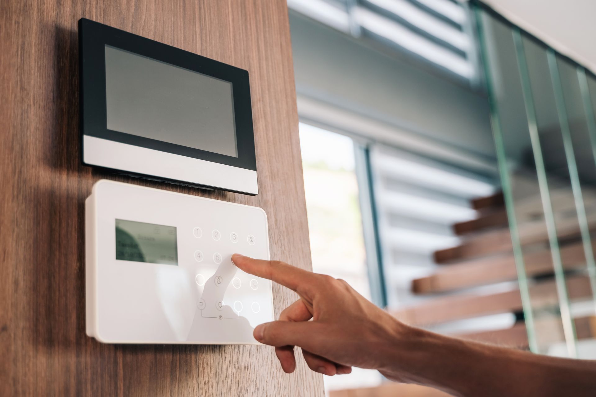 A person's hand pressing a button on a white security system control panel mounted on a wooden wall, with a black monitor above.