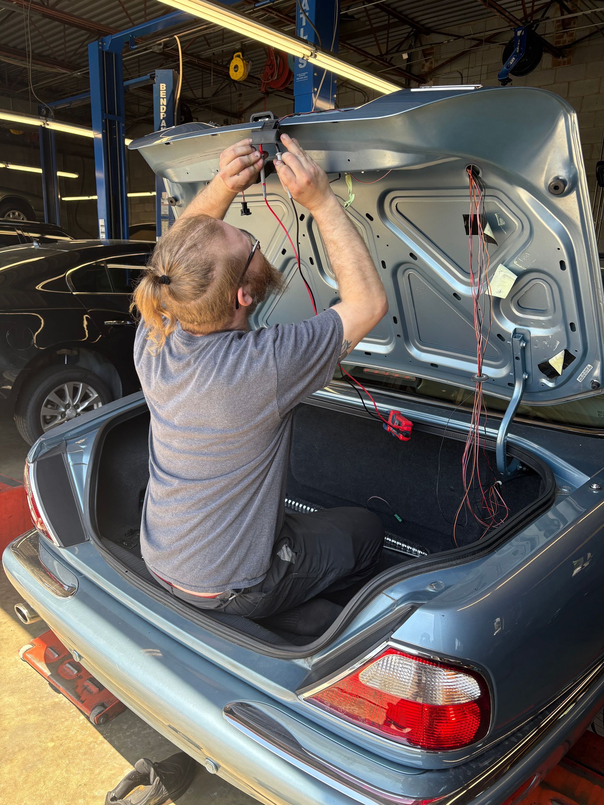 Person installing a trunk lid on a dark car in a garage, with the trunk open and tools nearby | Classic Import Auto Service