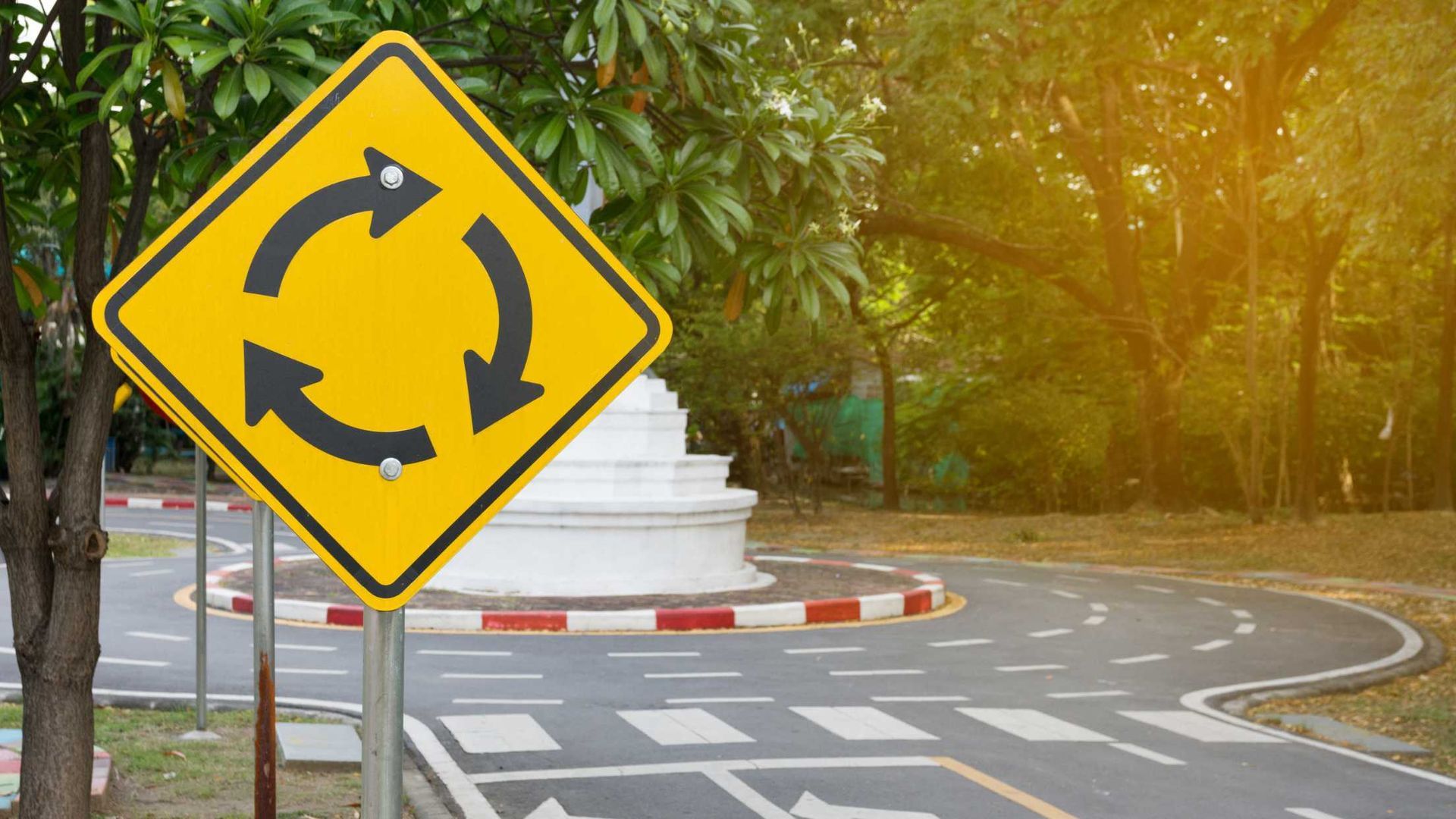 A yellow roundabout sign is sitting on the side of a road.