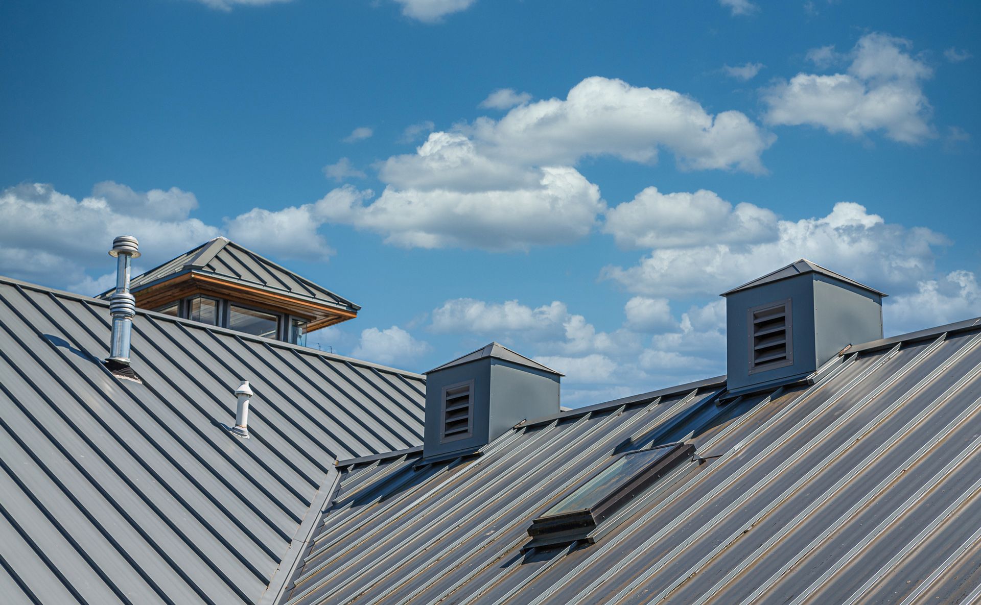 Gray metal roof with multiple chimneys against a blue sky with fluffy white clouds.