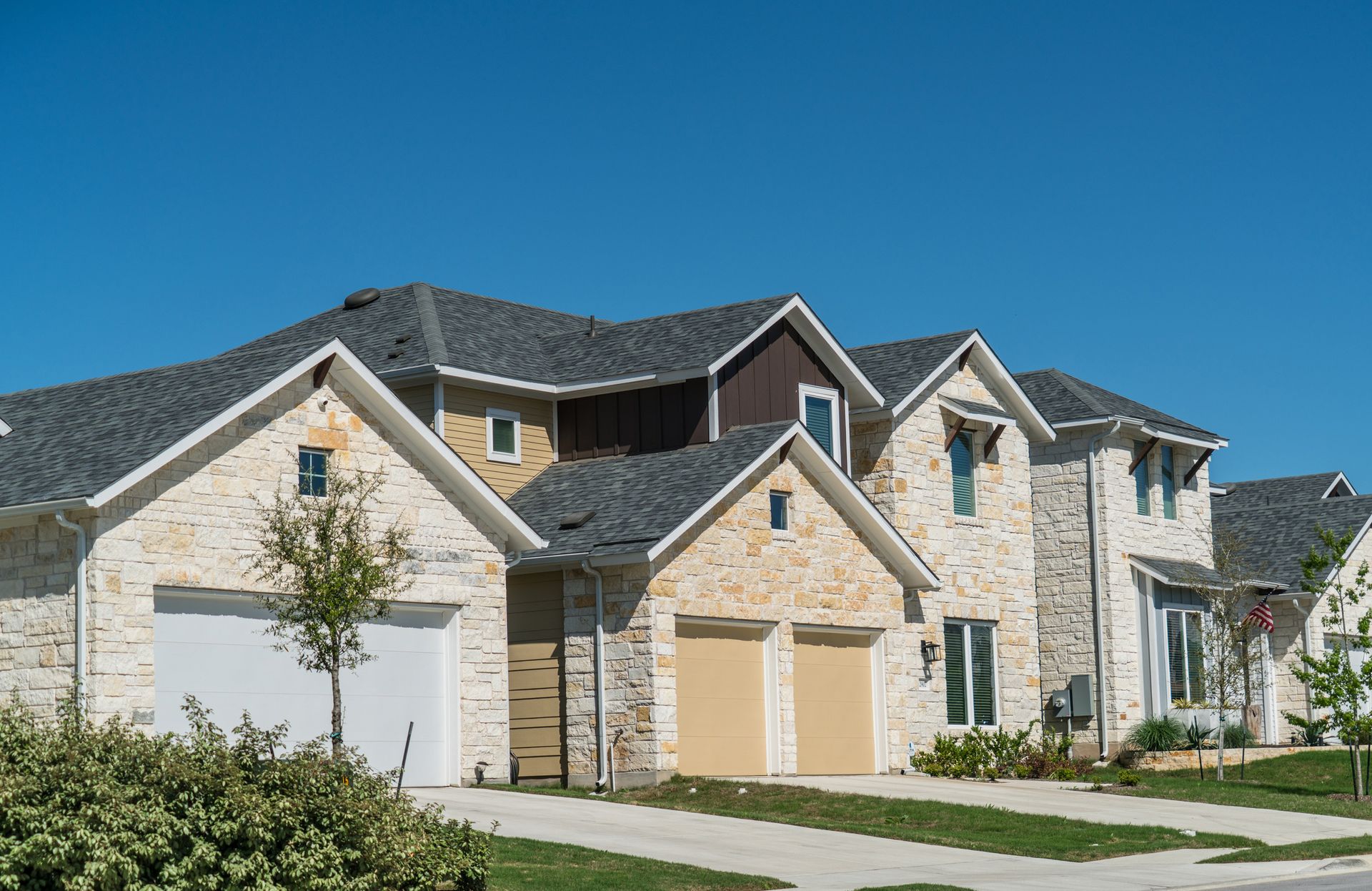 Row of houses with light-colored stone exteriors, white garage doors, and gray roofs under a blue sky.