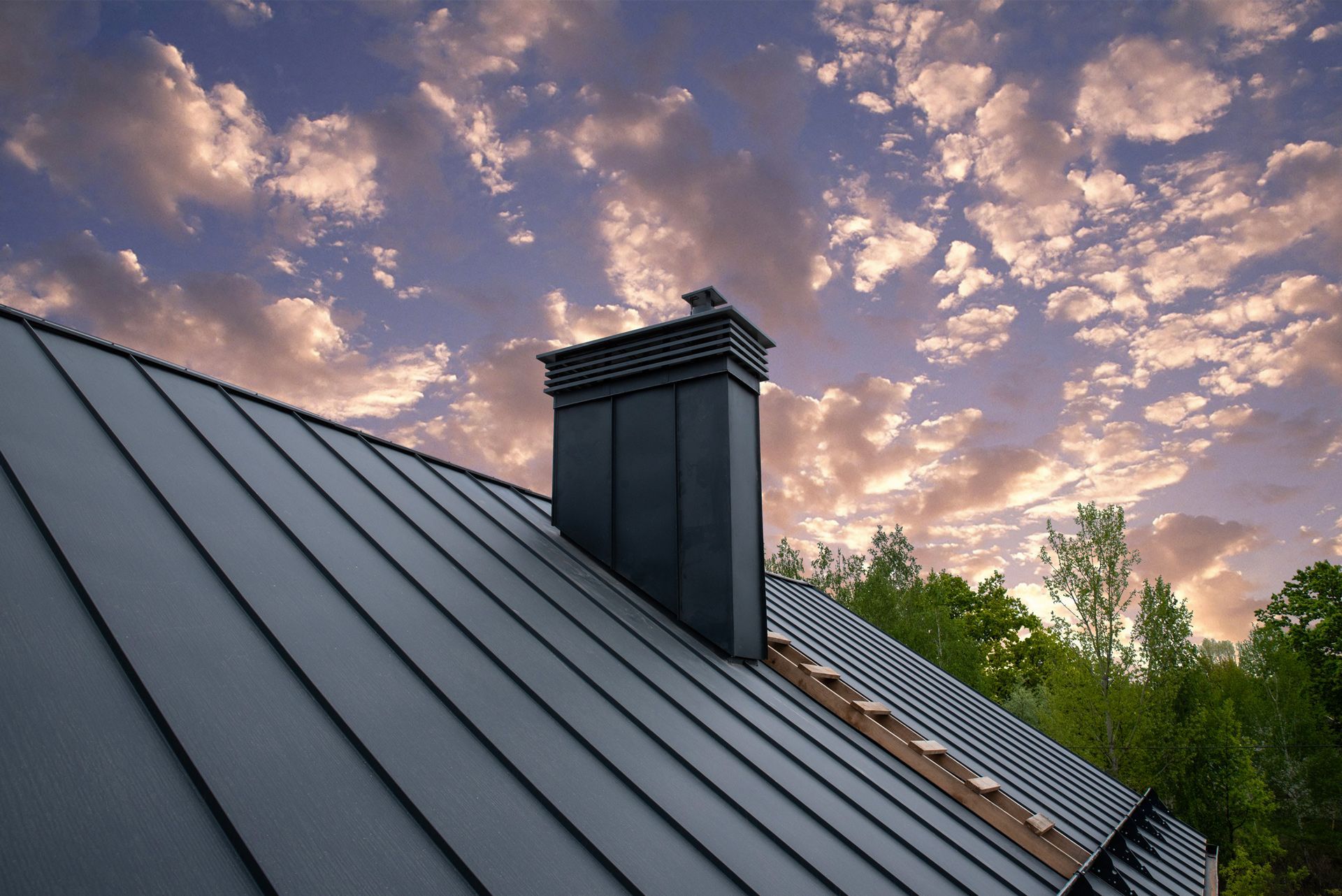 Dark metal roof with chimney against a sunset sky.