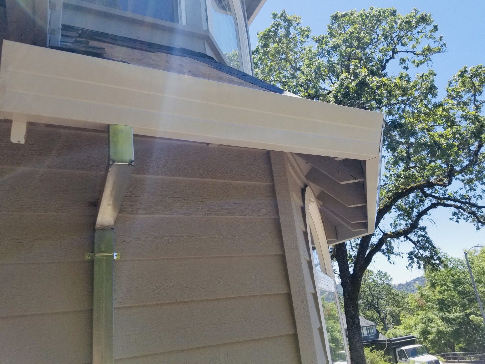 Side of a beige house with white trim and green supports, viewed outdoors beneath a blue sky and trees.