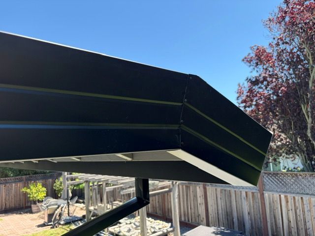 Black metal patio awning over a backyard with wooden fence and blue sky