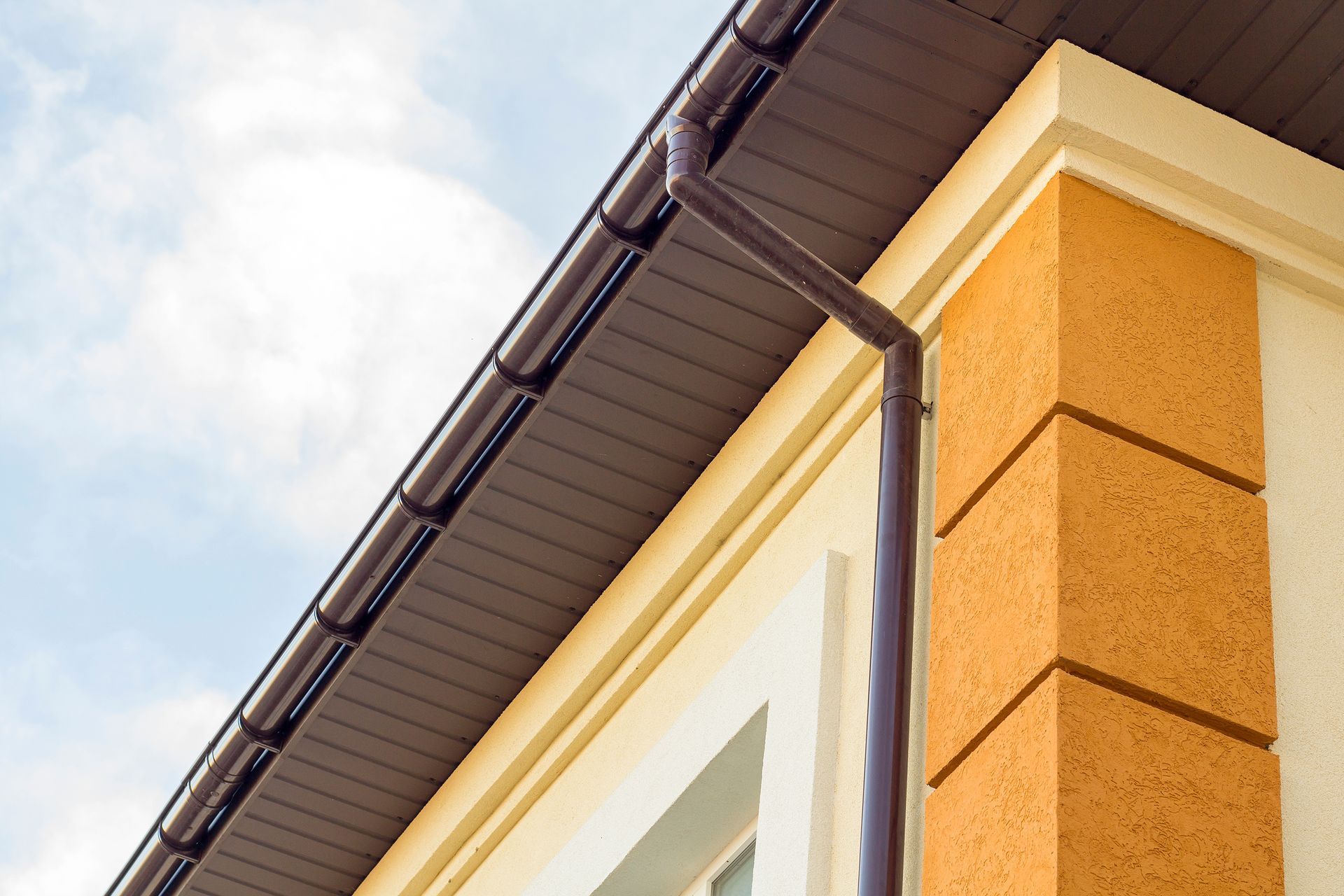 Close-up of a house eave with white trim, gray gutter, and orange stucco wall against a cloudy sky