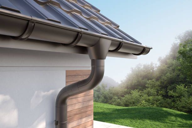 Rain gutter and downspout on a house roof beside a green yard and trees