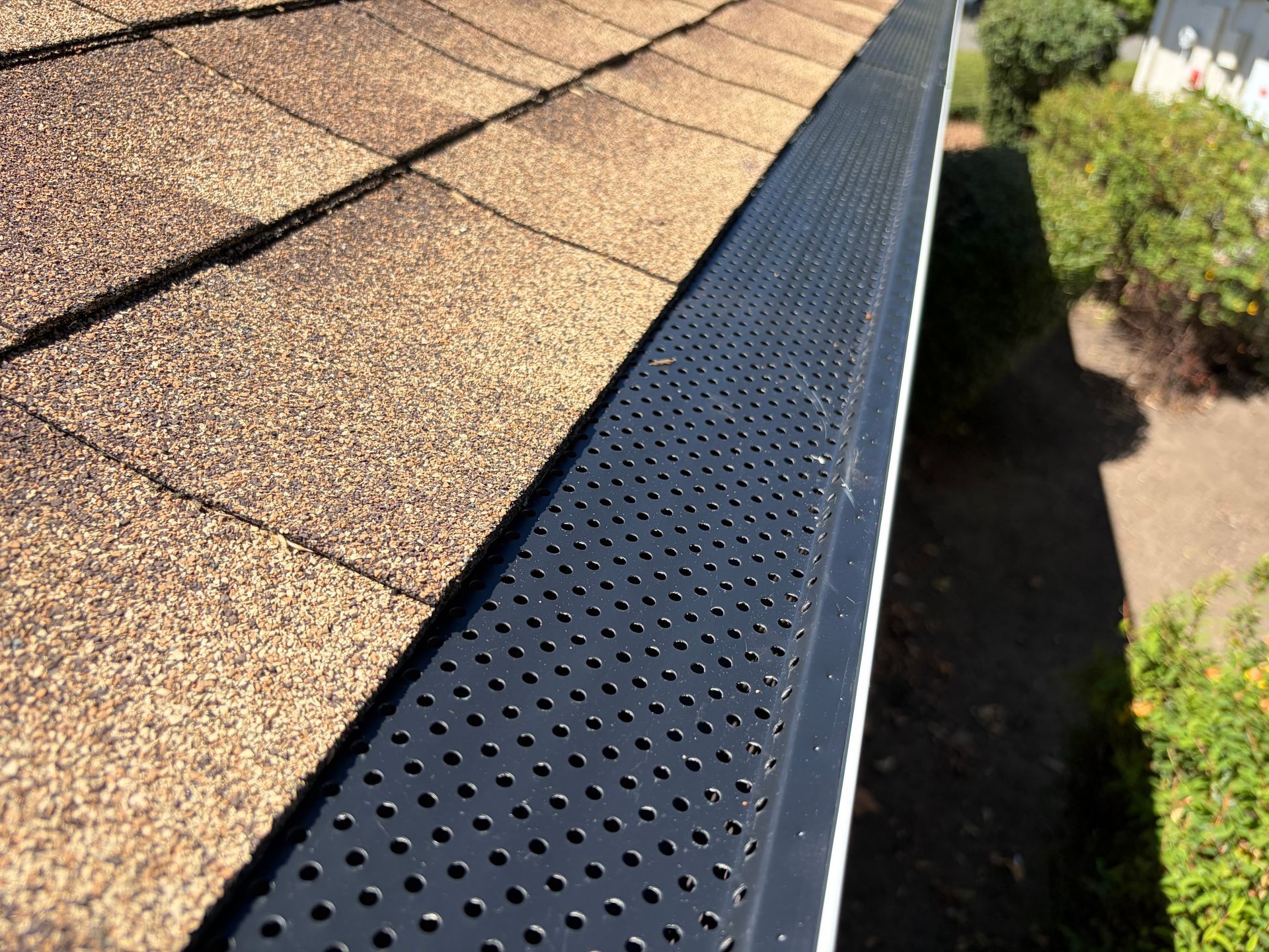 Roof gutter with black leaf guard beside tan shingles, viewed along the roof edge Roof gutter with black leaf guard beside tan shingles, viewed along the roof edge