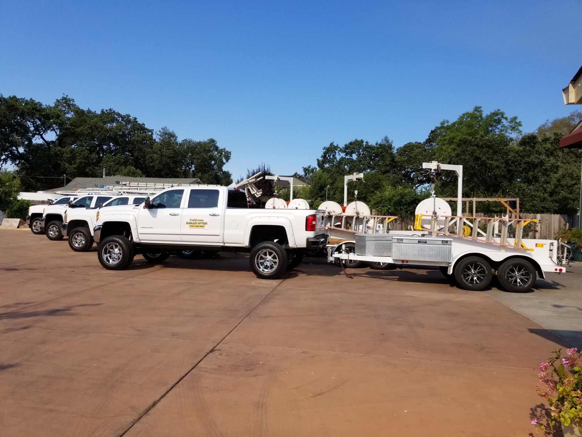 White dual-axle utility trailer with reel parked on a street near houses and trees under construction.