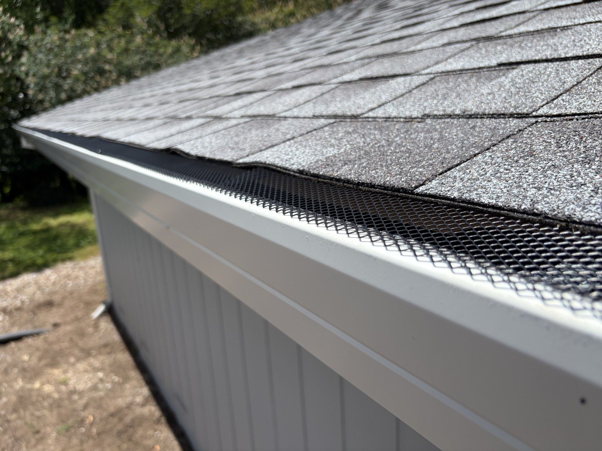 Close-up of a gray shingled roof with black leaf guard along the gutter and white siding below