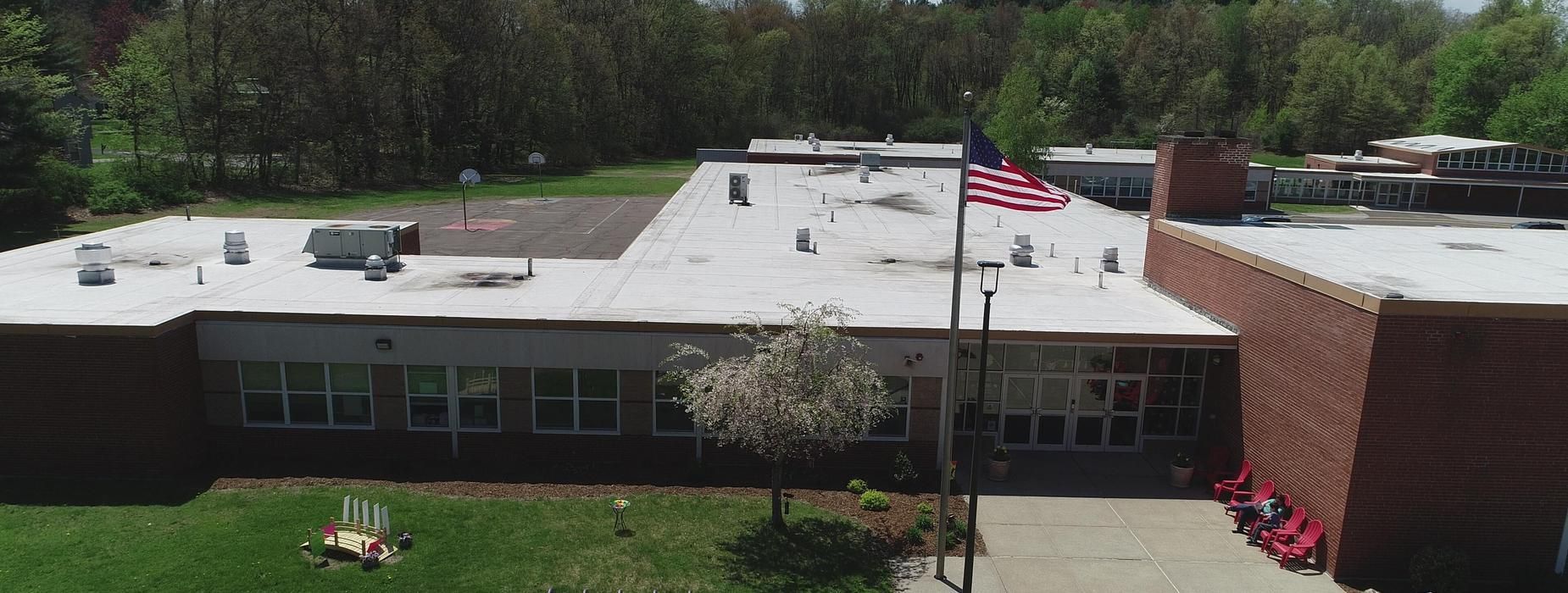 School building with American flag waving in the wind; surrounded by trees and a green lawn.