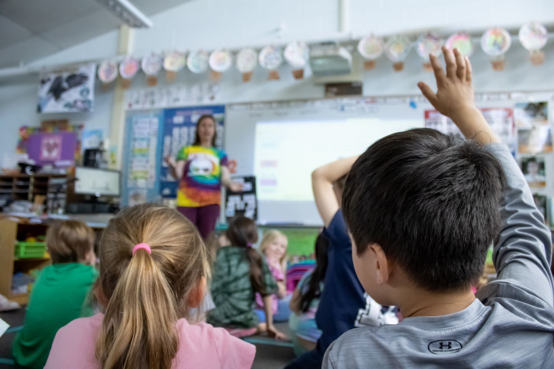 Students in a classroom, one with raised hand. Teacher at the projector screen.
