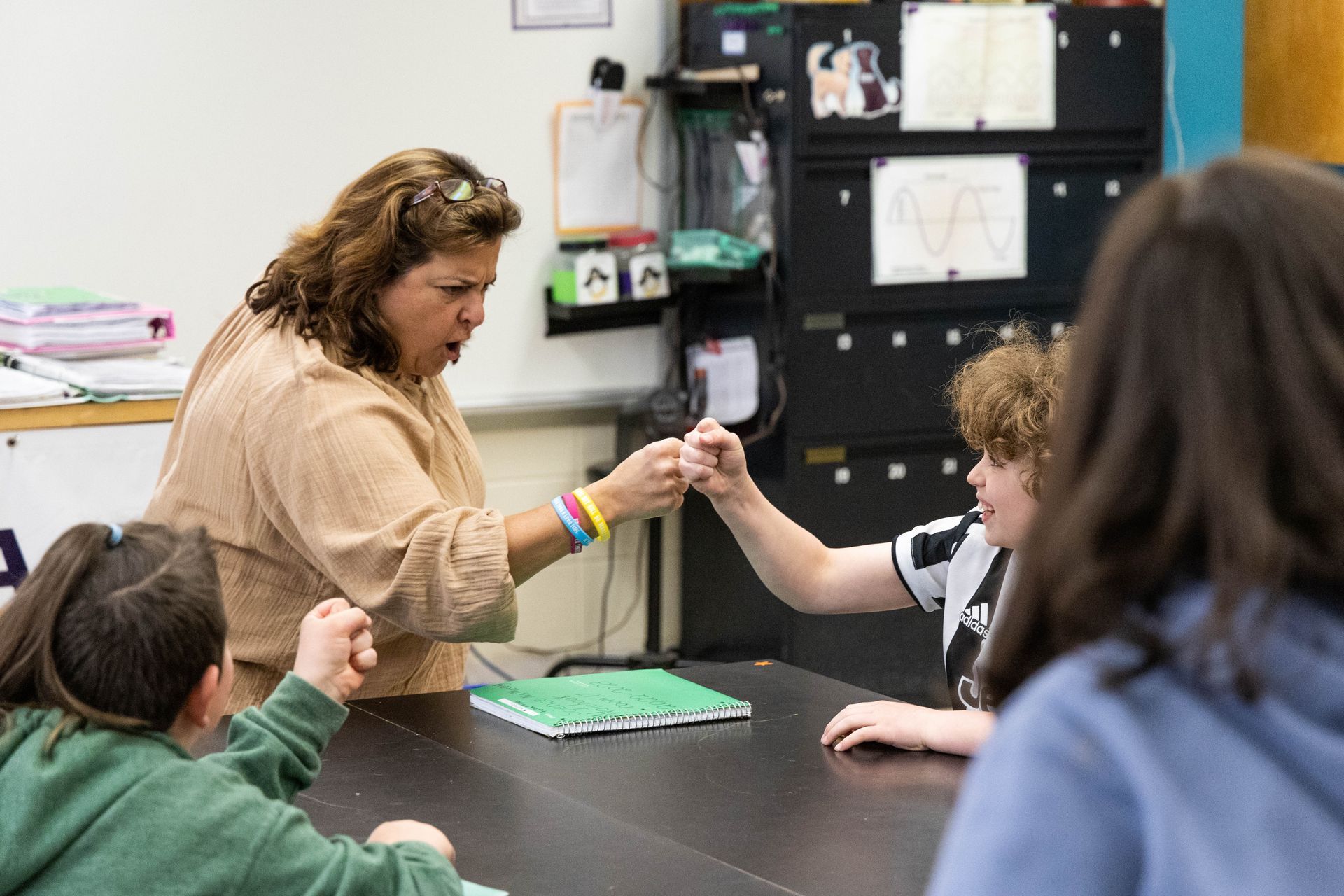 Teacher fist-bumps a student over a table; two other students watch in a classroom.