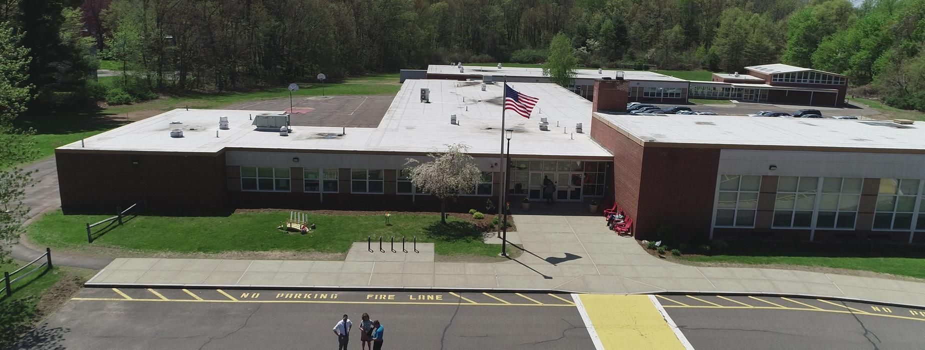 An aerial view of a brick schoolhouse with a flag, some trees, and people walking on the sidewalk.